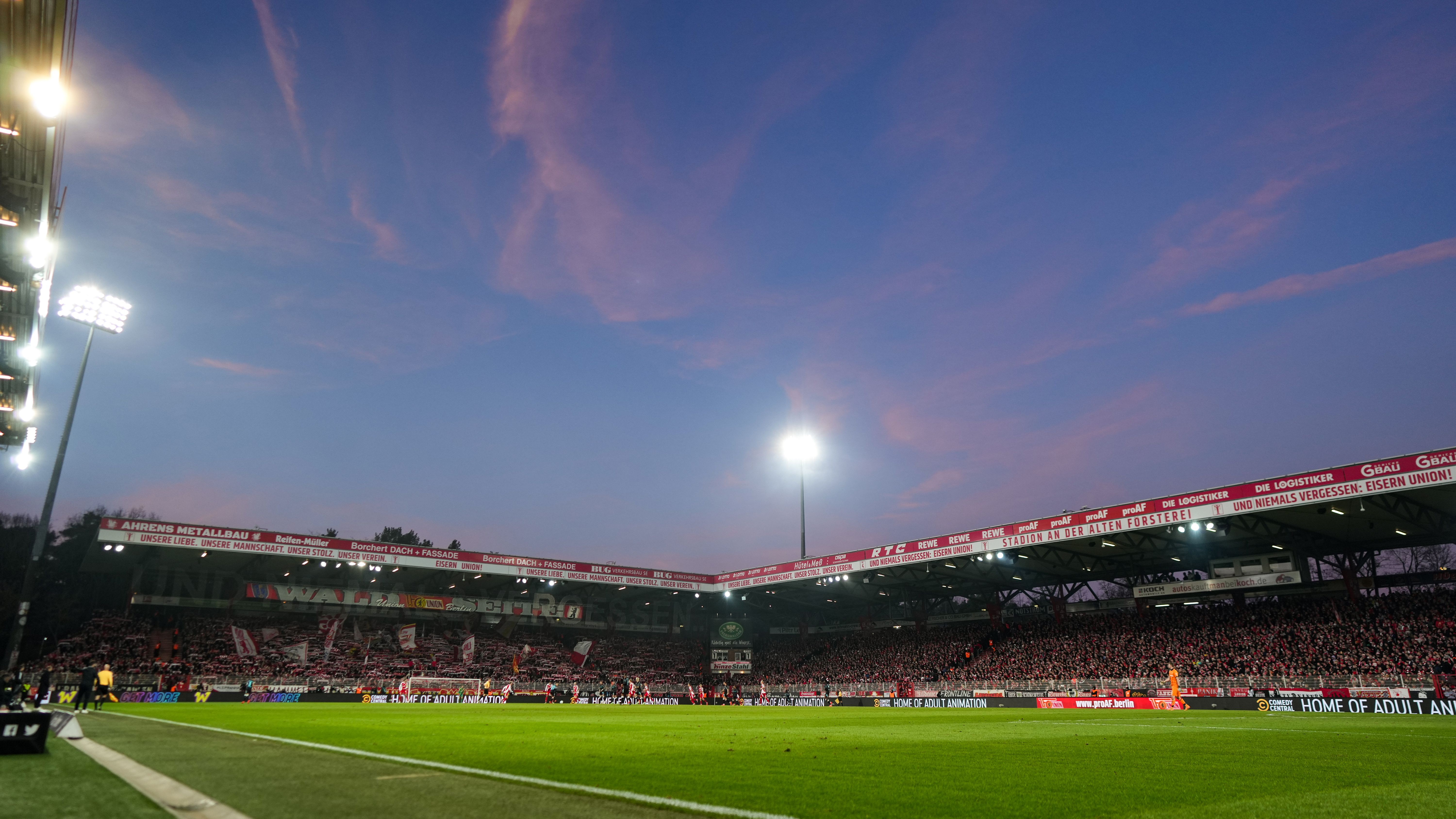 Fußballspiel im Stadion bei Dämmerung, überfüllte Tribüne und farbenfroher Himmel im Hintergrund.