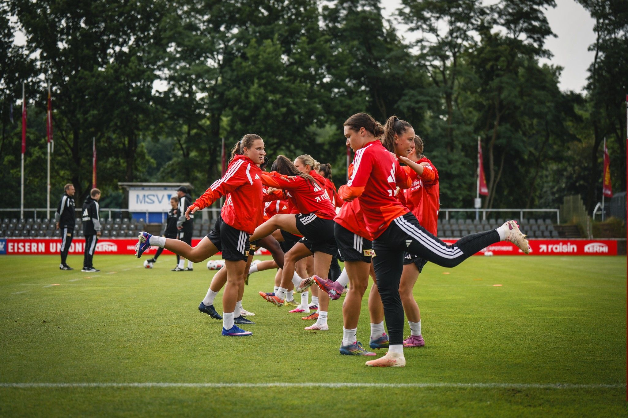 Frauenfußballmannschaft in roten Trainingsjacken, die in einer Reihe Beinübungen auf dem Spielfeld ausführen.