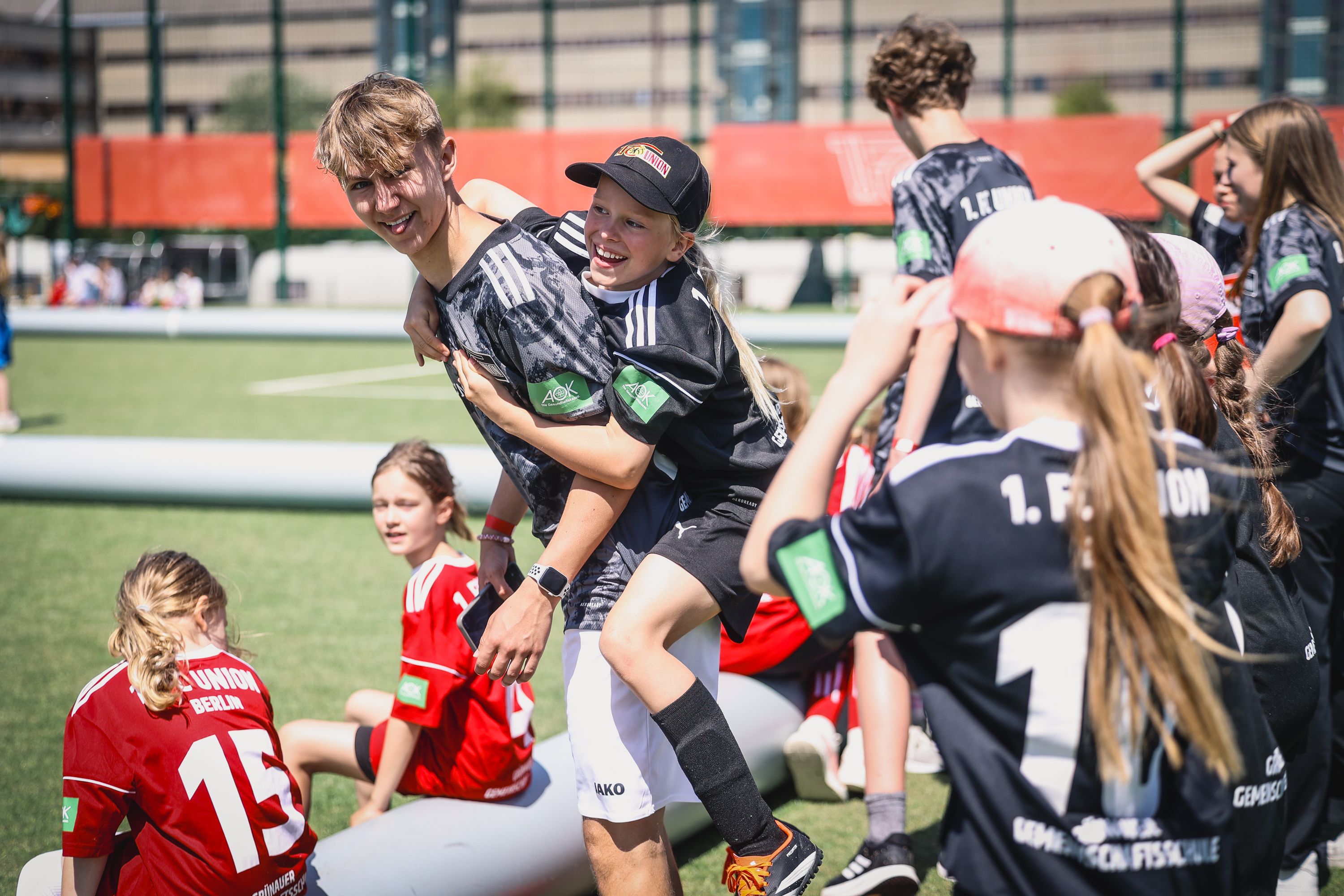 Ein Junge trägt einen anderen Jungen auf dem Rücken, während mehrere Kinder in Fußballtrikots auf dem Rasen spielen und lachen.