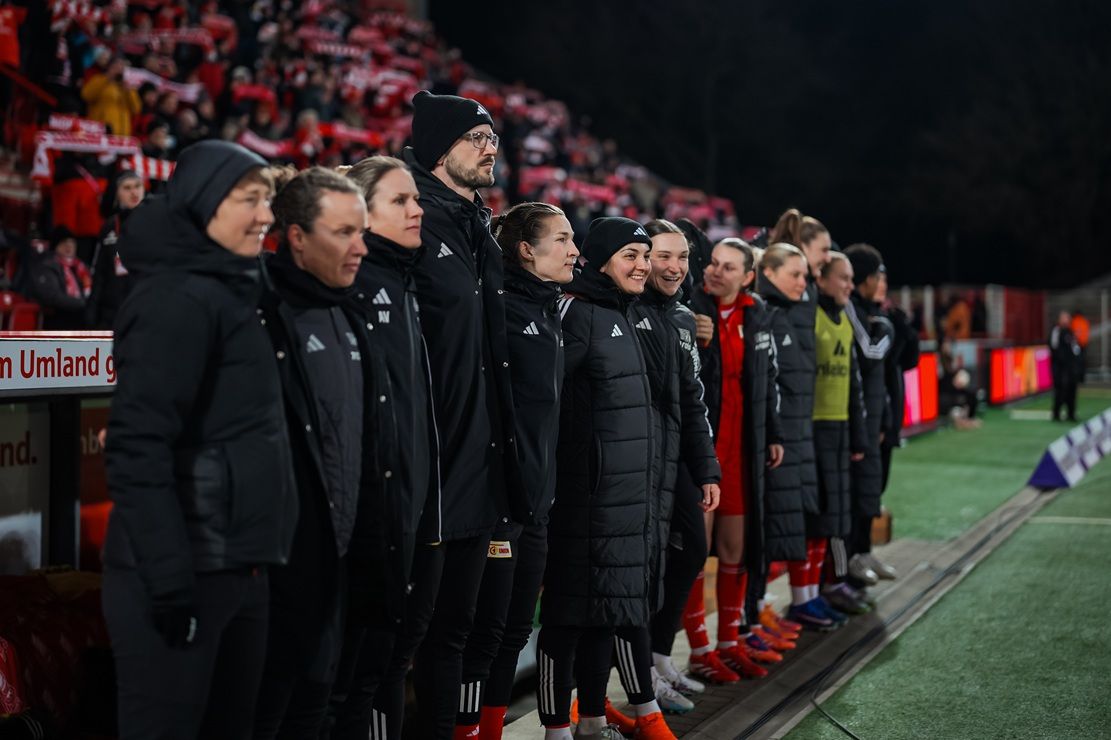 Die Bank der Spielerinnen steht auf der Seitenlinie eines Fußballfeldes, während sie ein Spiel beobachten.