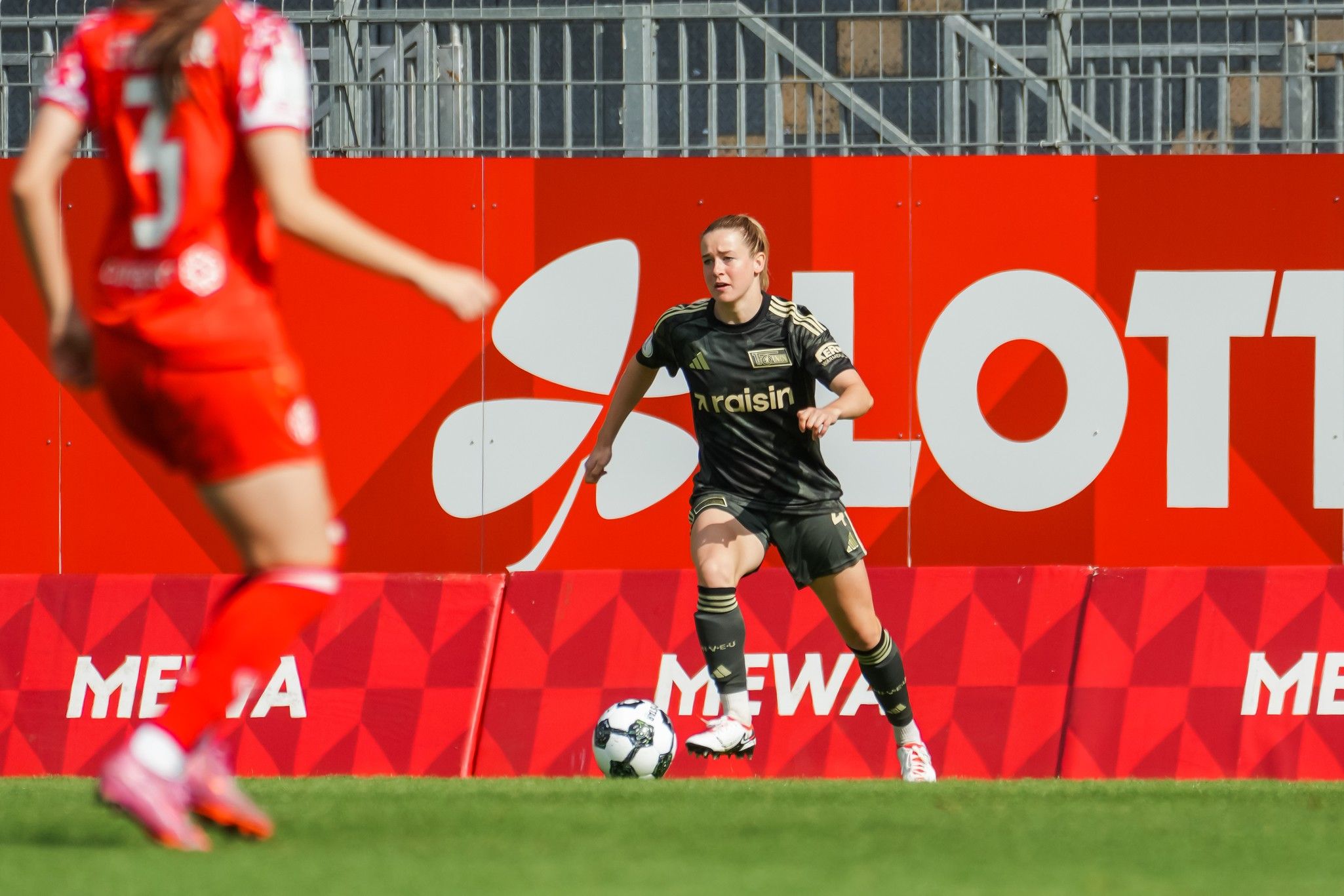 Eine Spielerin in schwarzer Sportkleidung dribbelt den Ball auf dem Feld, während eine Gegnerin in roter Kleidung im Hintergrund zu sehen ist.