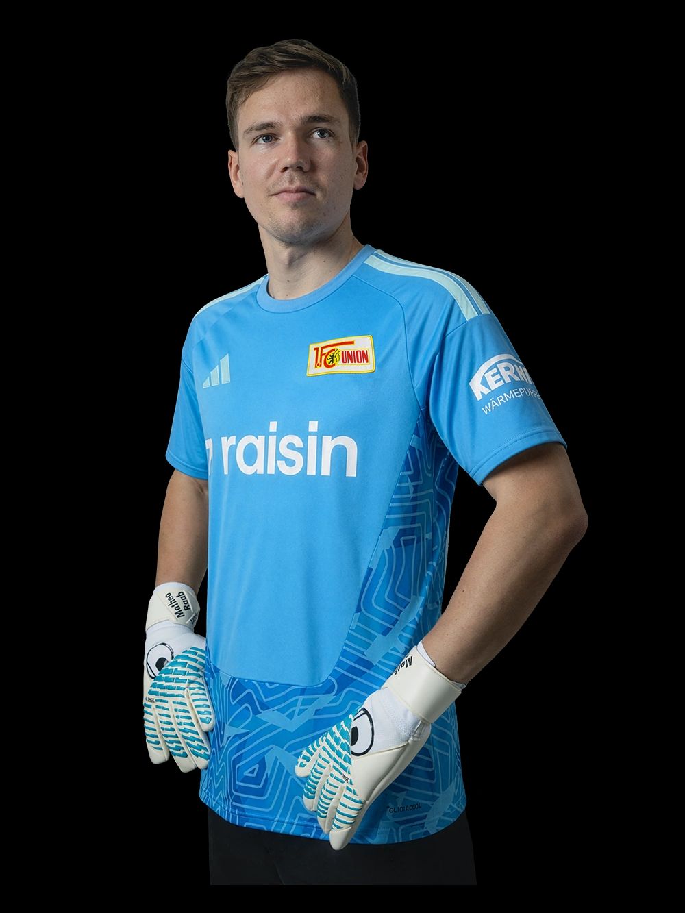 Ein junger Torwart in einem blauen Trikot mit grafischen Mustern und weißen Handschuhen, posiert vor schwarzem Hintergrund.