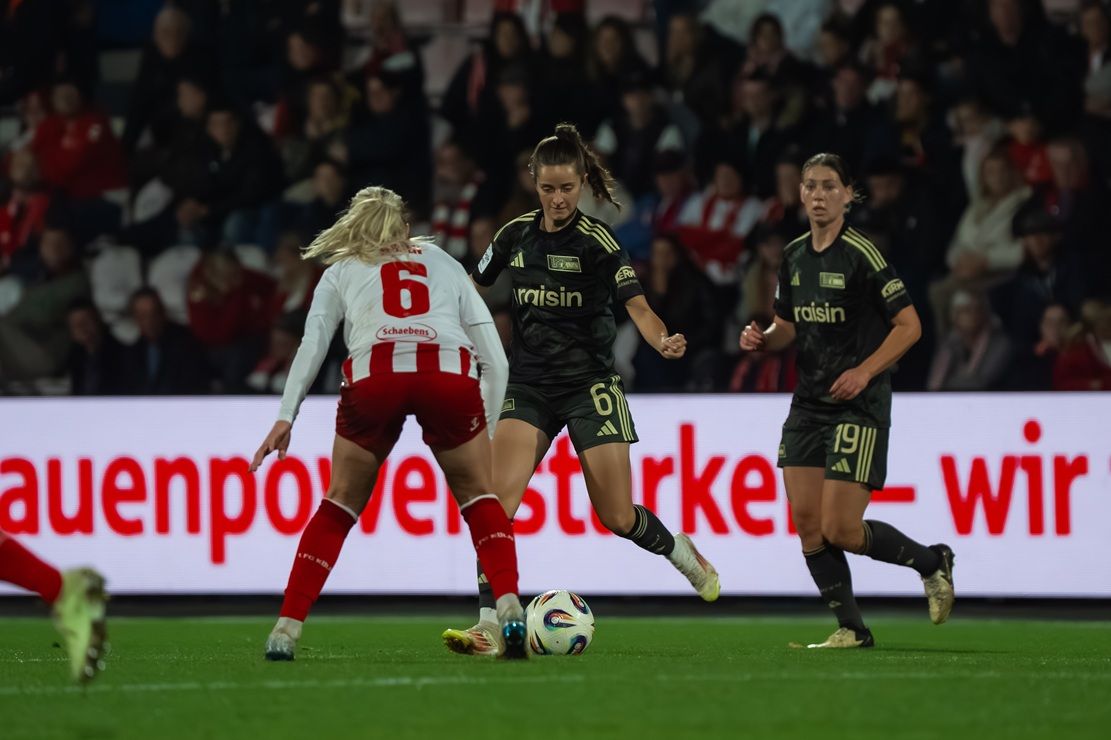 Zwei Fußballspielerinnen in schwarzen und roten Trikots antreten auf einem Spielfeld, während eine Spielerin den Ball kontrolliert.