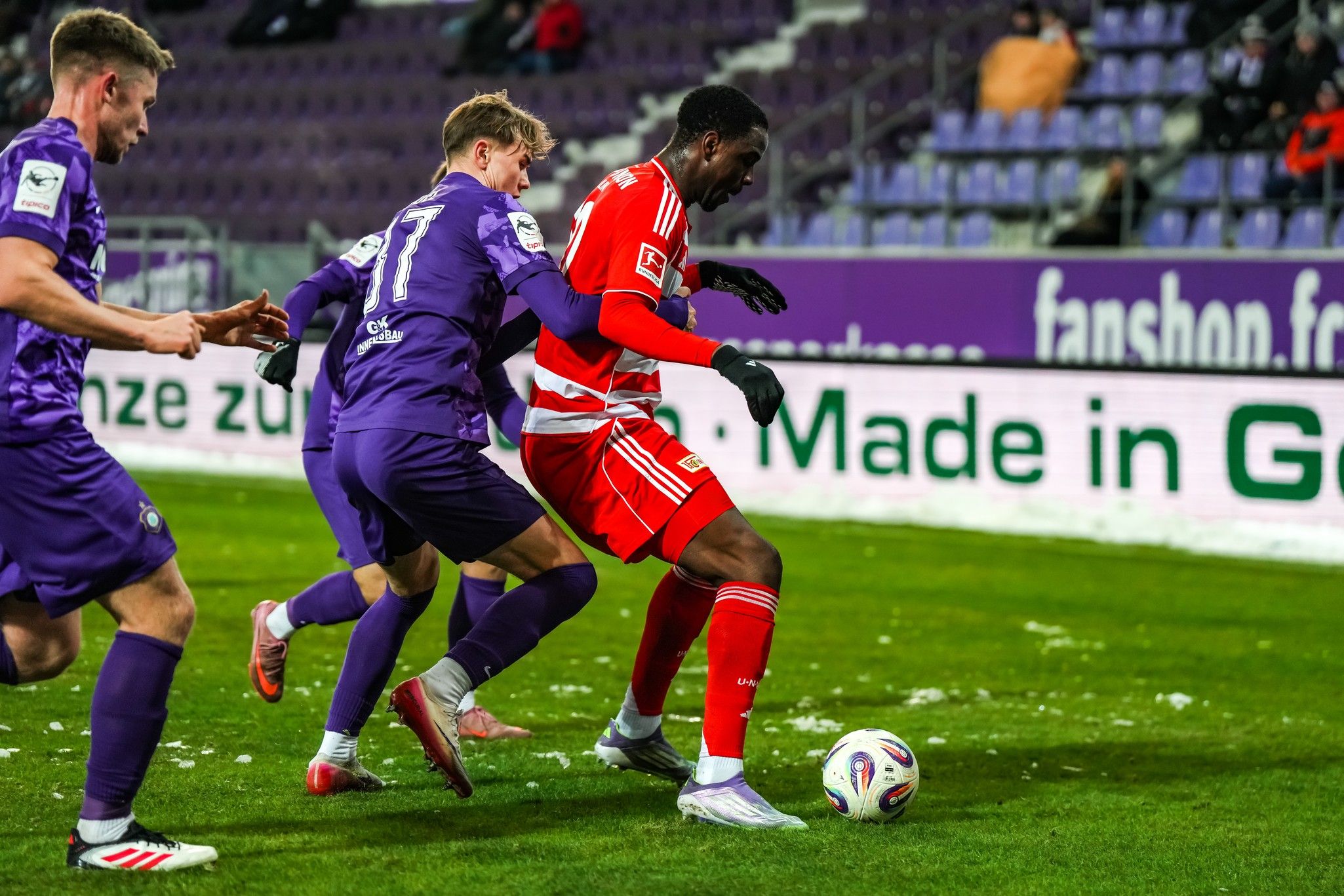 Zwei Fußballspieler in lila und rot kämpfen um den Ball auf einem Platz mit schneebedecktem Terrain.