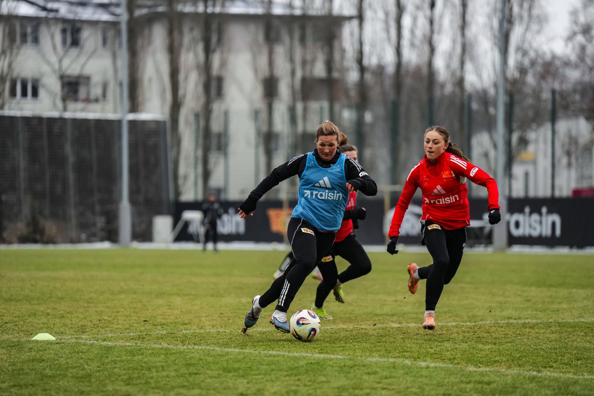 Zwei Fußballspielerinnen sprinten über einen Rasenplatz, eine trägt ein blaues Trainingsshirt, die andere rot.