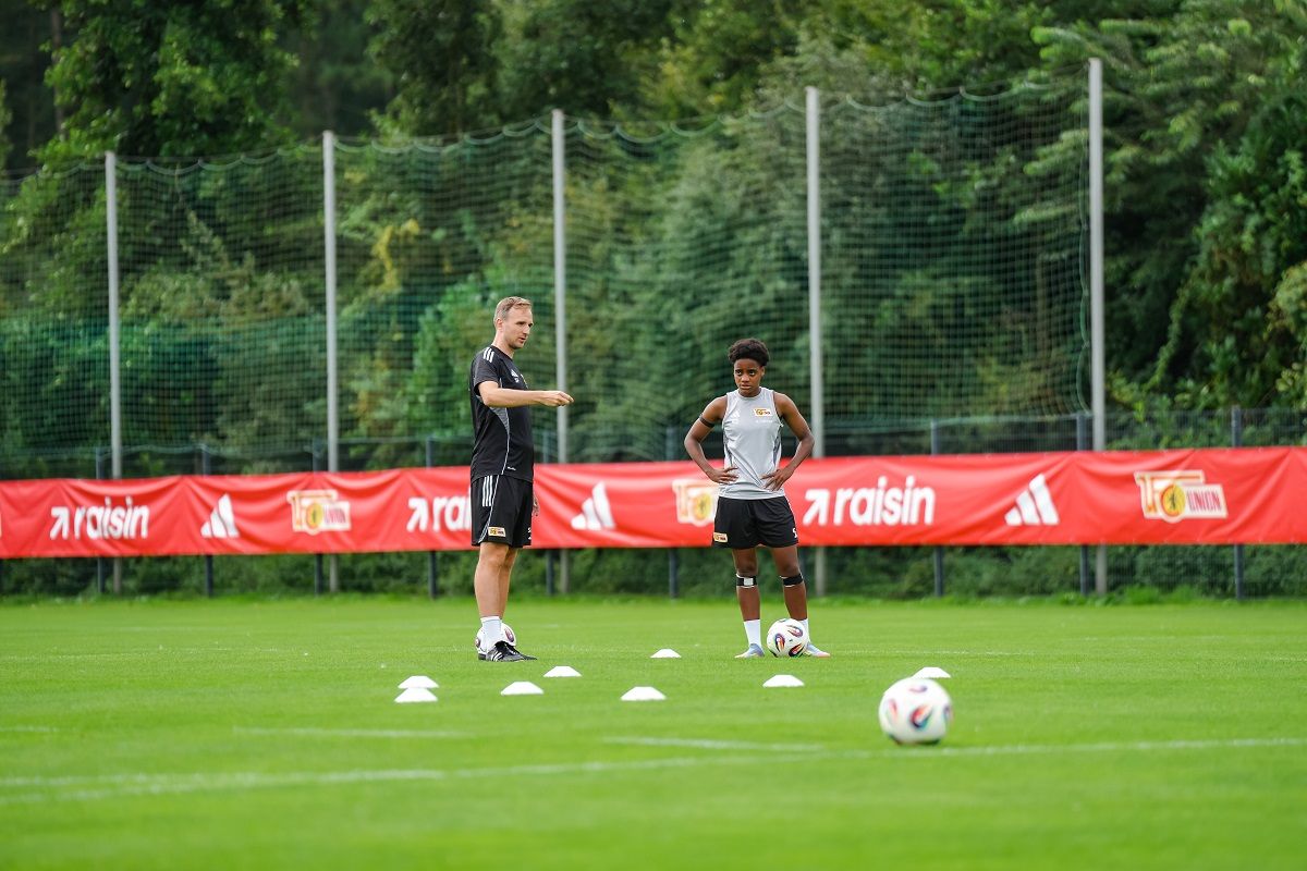 Ein Trainer gibt Anweisungen an einen Spieler während eines Fußballtrainings auf einem Rasenplatz mit Markierungen.