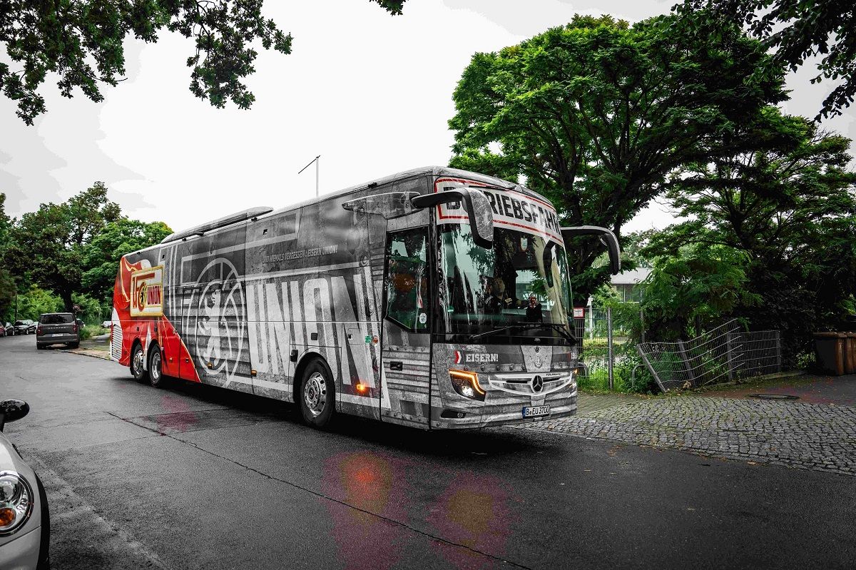 Ein großer, grauer Bus mit Werbung für "Union" parkt an einer Straße, umgeben von Bäumen und einem Wohngebiet.