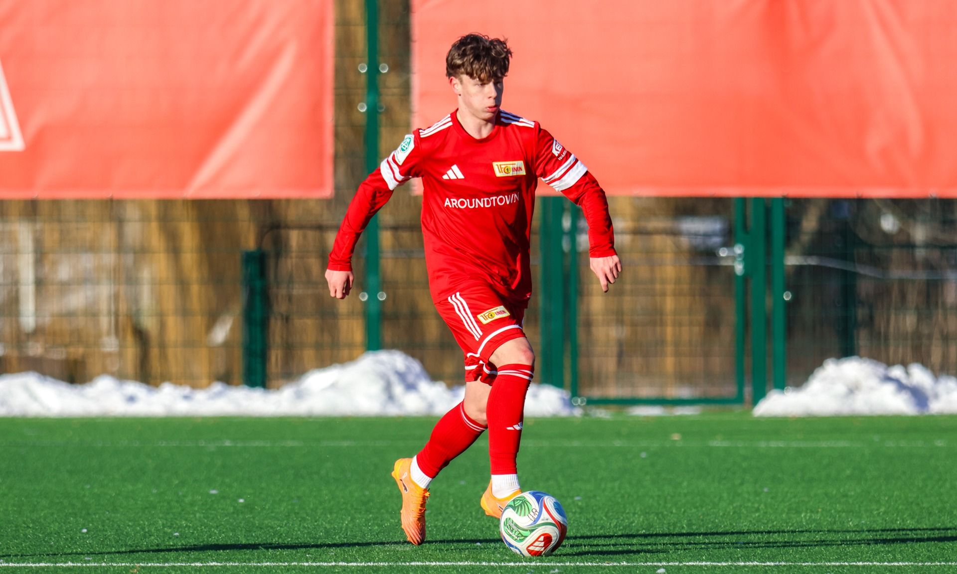 Ein junger Fußballspieler in rotem Trikot verirrt einen Ball auf einem grünen Kunstrasenfeld.