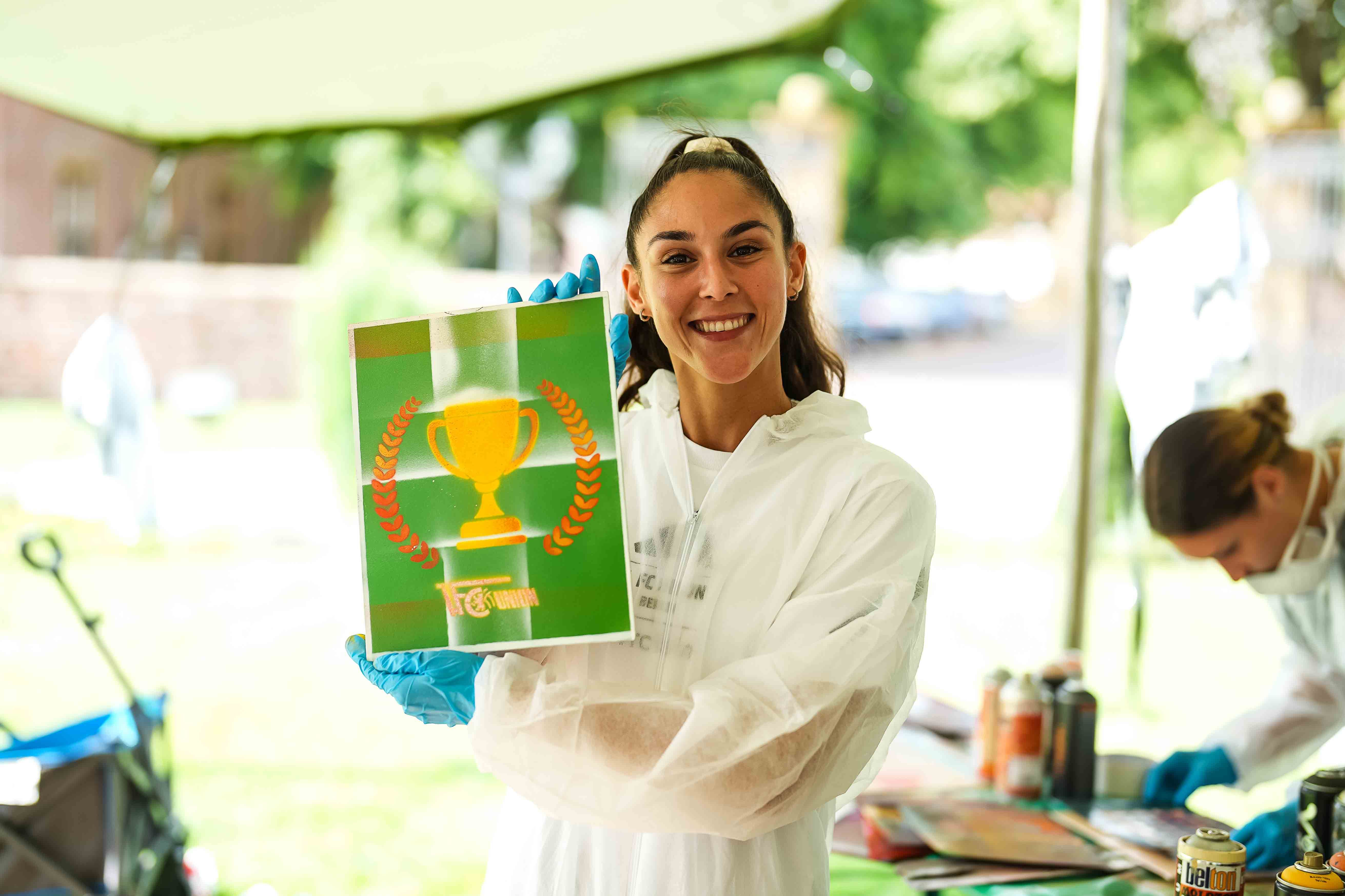 Eine lächelnde Frau in Schutzkleidung hält ein grünes Plakat mit einem Pokal und Lorbeerkranz.