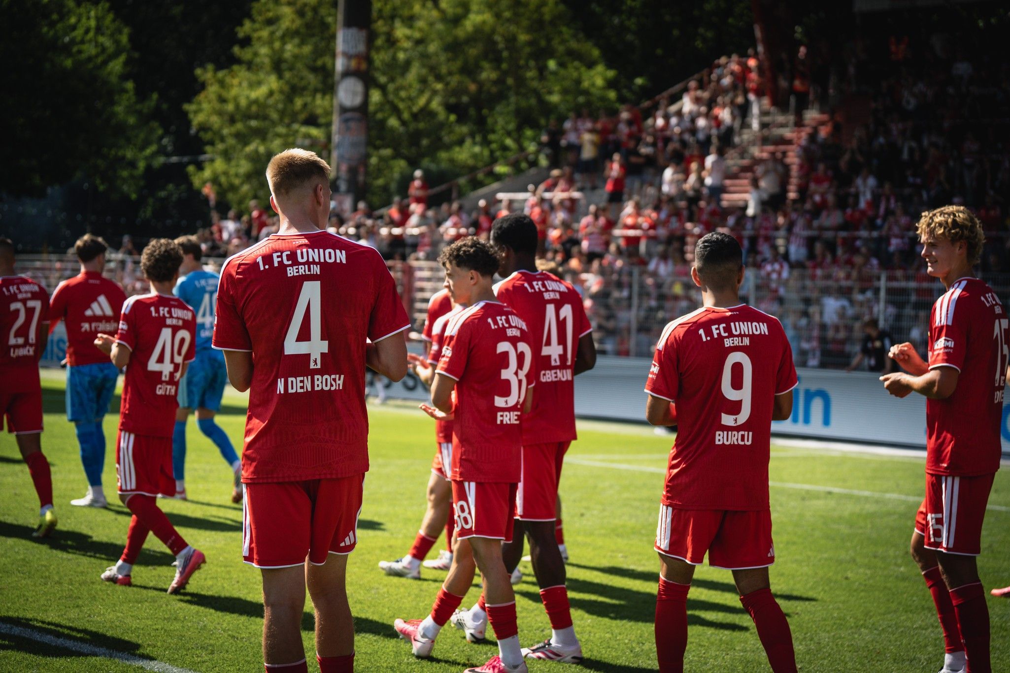 Spieler des 1. FC Union Berlin in roten Trikots stehen auf dem Spielfeld, im Hintergrund eine Zuschauertribüne.