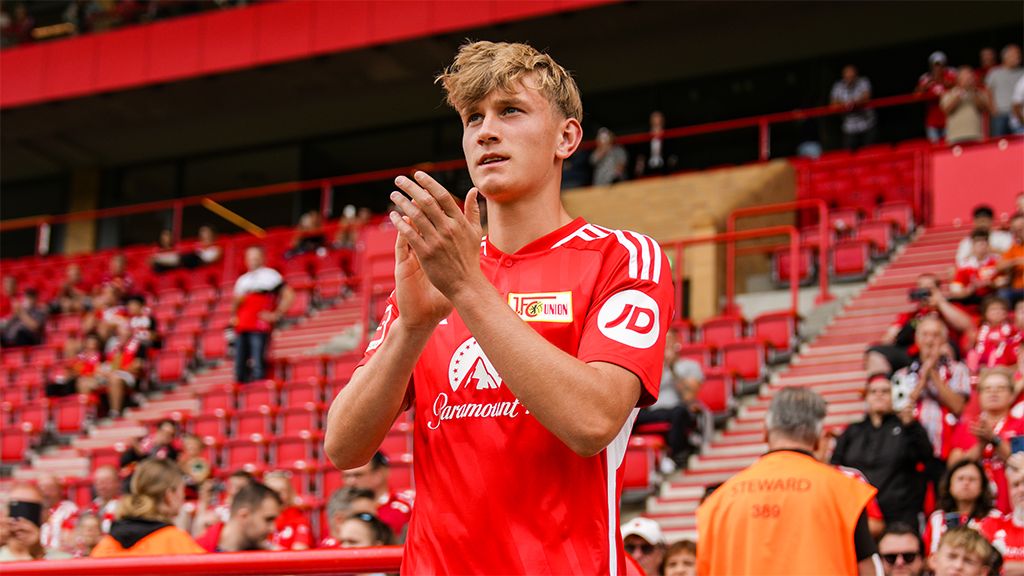 Young football player in a red jersey claps his hands, surrounded by cheering fans in a stadium.