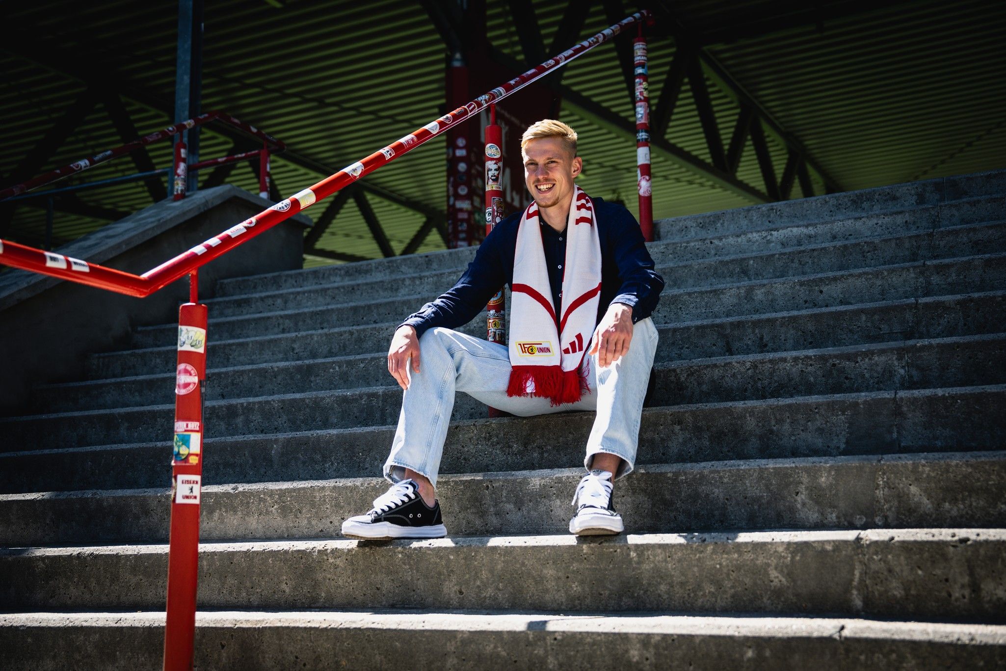 Ein lächelnder junger Mann sitzt auf einer Treppe in einem Stadion, trägt eine Fussballschal und hat legere Kleidung an.