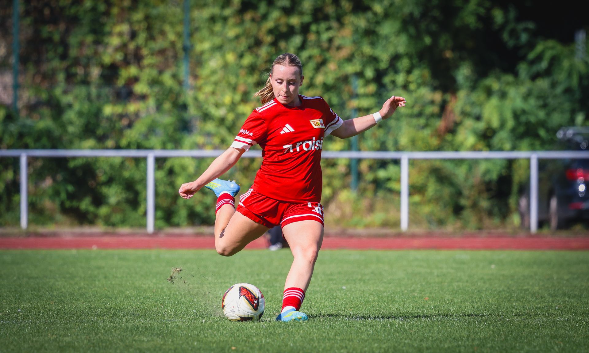 Eine Fußballspielerin in rotem Trikot tritt einen Ball auf einem Sportplatz. Im Hintergrund sind Bäume und eine Tribüne zu sehen.