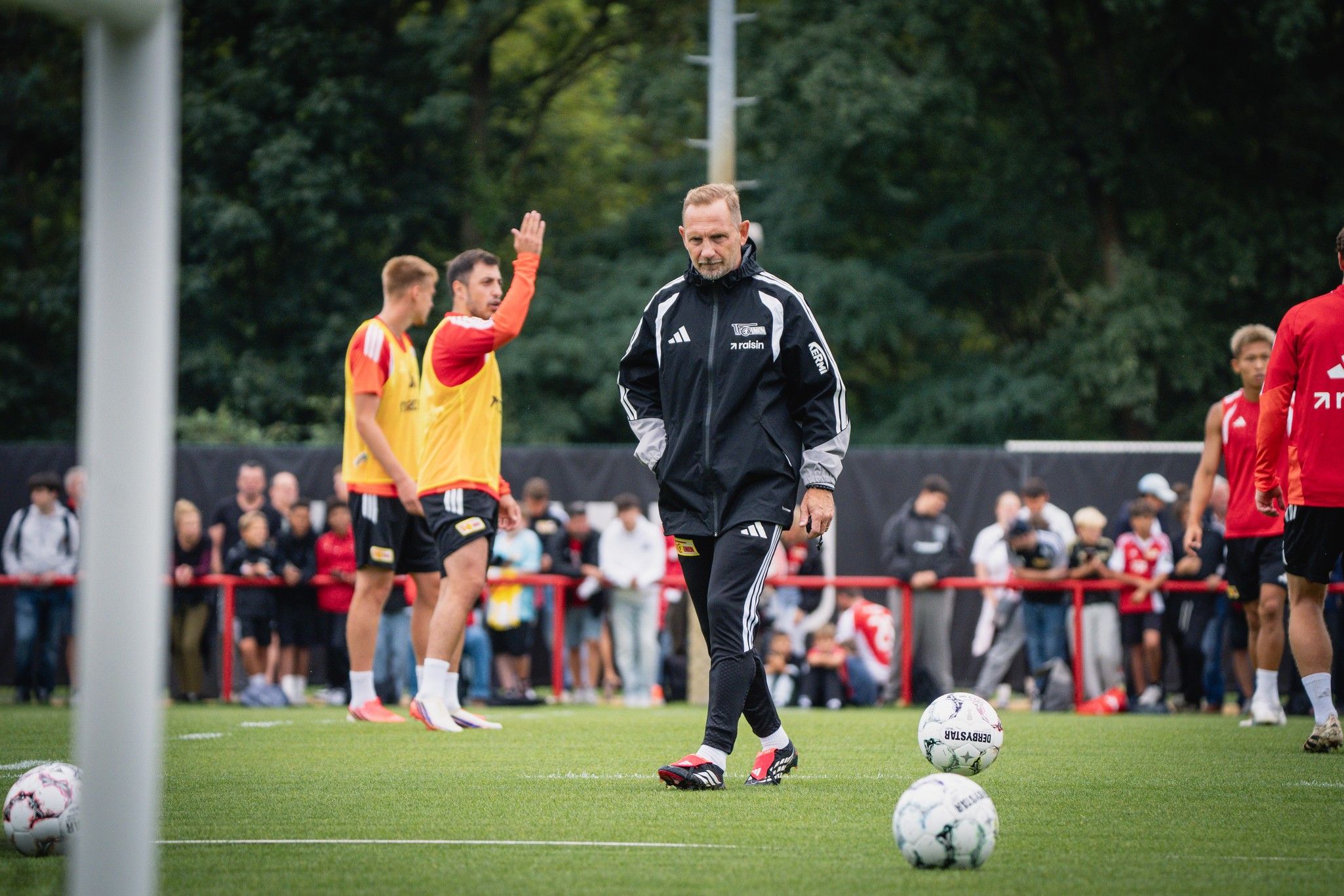 Ein Trainer beobachtet seine Spieler während des Trainings auf einem Fußballplatz, umgeben von Fans.