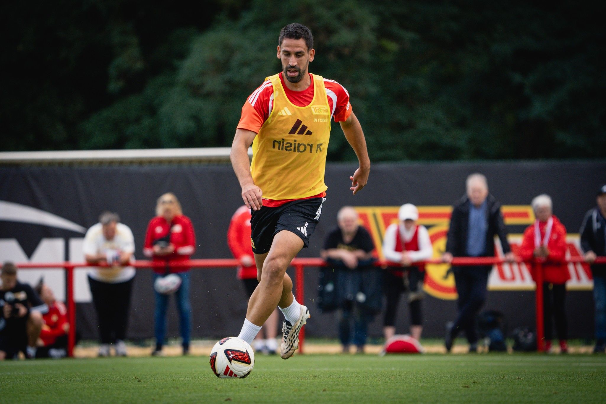 Ein Fußballspieler in einem Trainingsshirt läuft auf einem Spielfeld und dribbelt einen Ball. Zuschauer sind im Hintergrund zu sehen.
