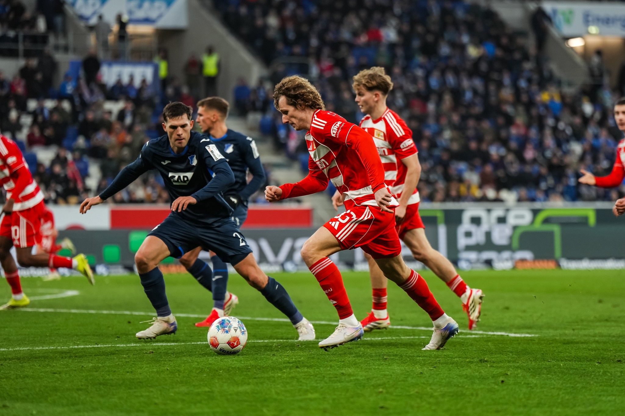 Ein Fußballspiel mit Spielern in roten und blauen Trikots, die einen Ball im Stadion verfolgen. Fans sind im Hintergrund sichtbar.