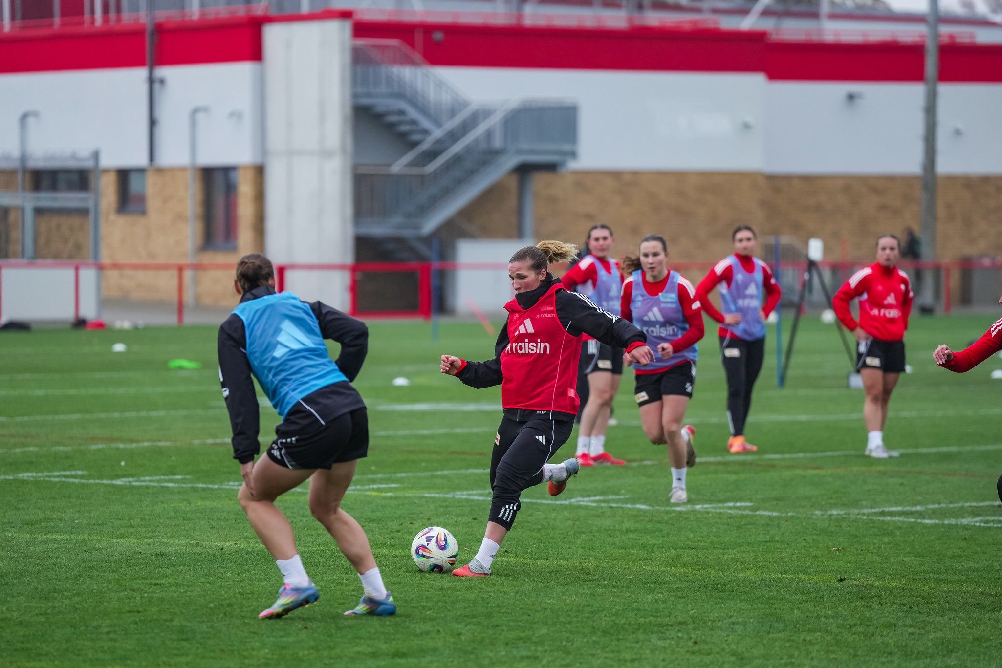 Frauenfußballmannschaft trainiert auf einem grünen Platz; Spielerinnen in roten und blauen Trikots bewegen sich während des Trainings.