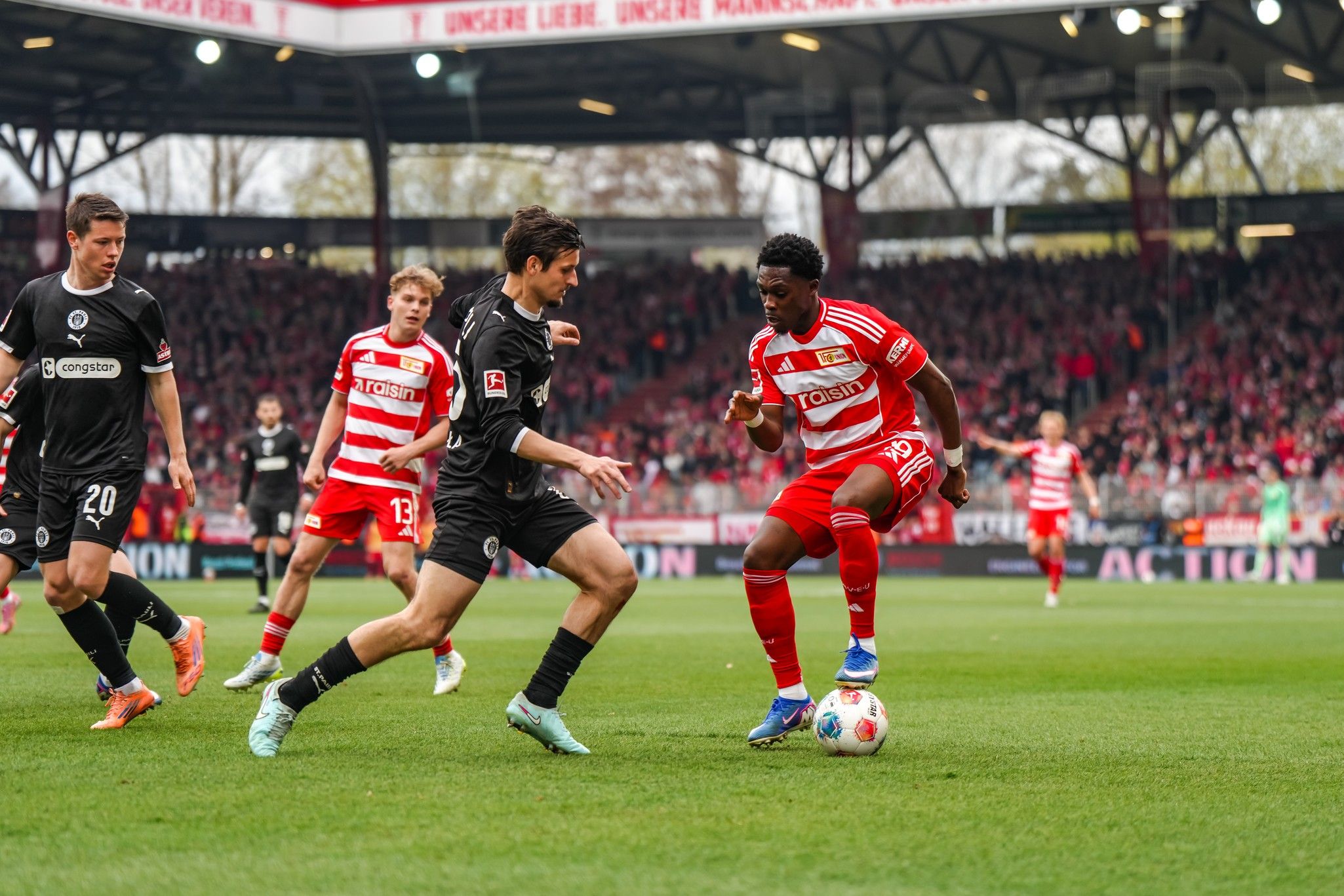 Zwei Fußballspieler in Aktion während eines Spiels, umgeben von Zuschauern im Stadion. Rot-weiße und schwarze Trikots sind sichtbar.