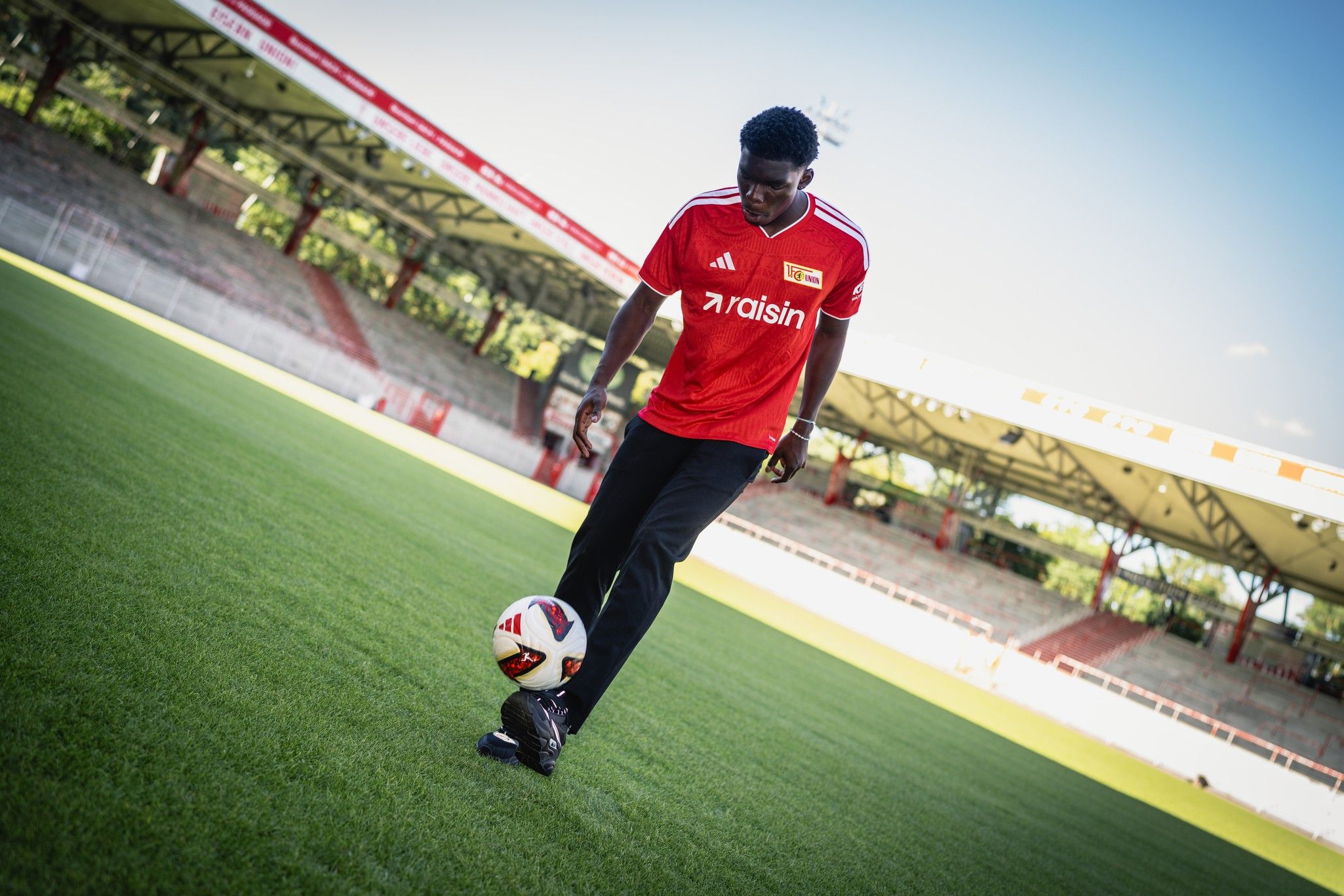 Ein Fußballspieler in rotem Trikot spielt mit einem Ball auf einem gepflegten Fußballfeld in einem Stadion.