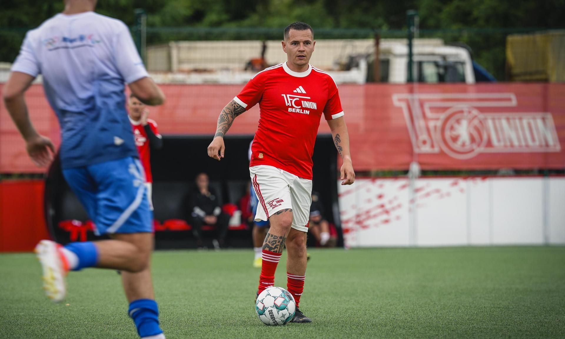Ein Fußballspieler in rotem Trikot dribbelt den Ball auf einem Kunstrasenplatz, während andere Spieler im Hintergrund aktiv sind.