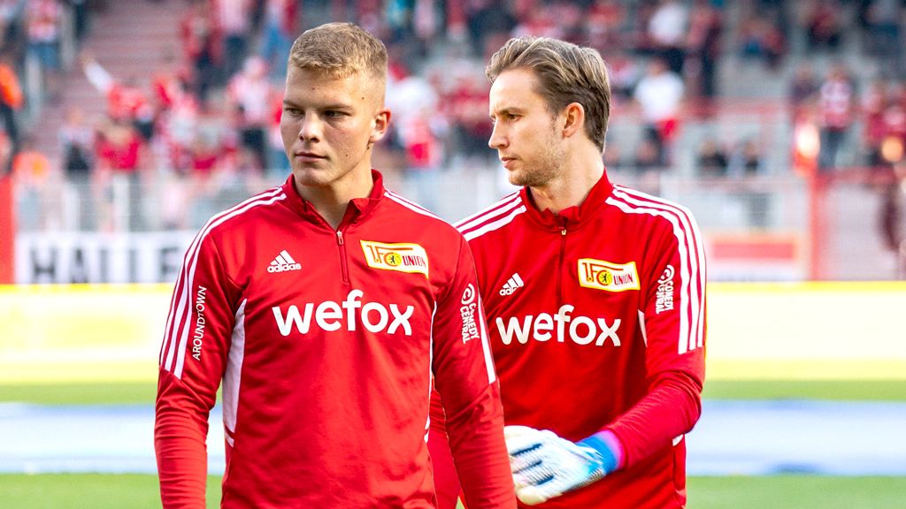 Zwei Fußballspieler in roten Trainingsanzügen mit dem Logo "wefox" betreten das Spielfeld.