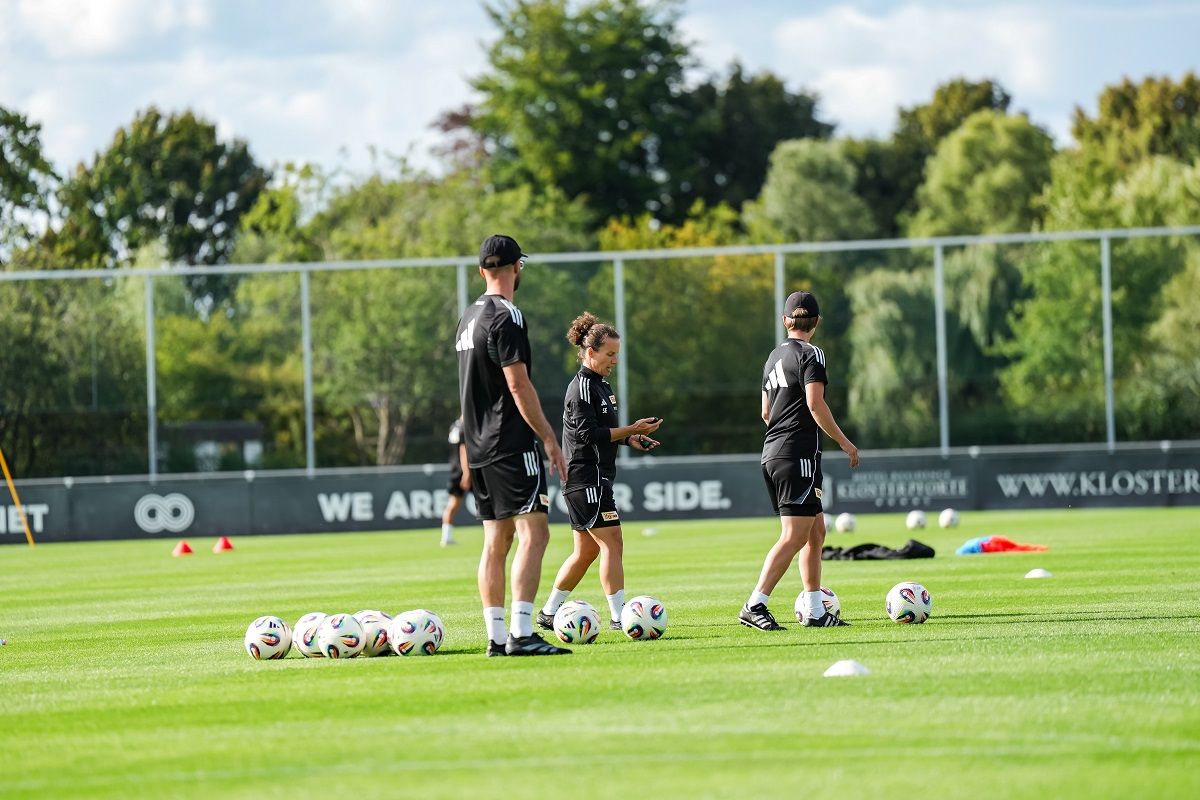 Drei Fußballspieler absolvieren ein Training auf einem grünen Platz, umgeben von Fußbällen und einem klaren Himmel.