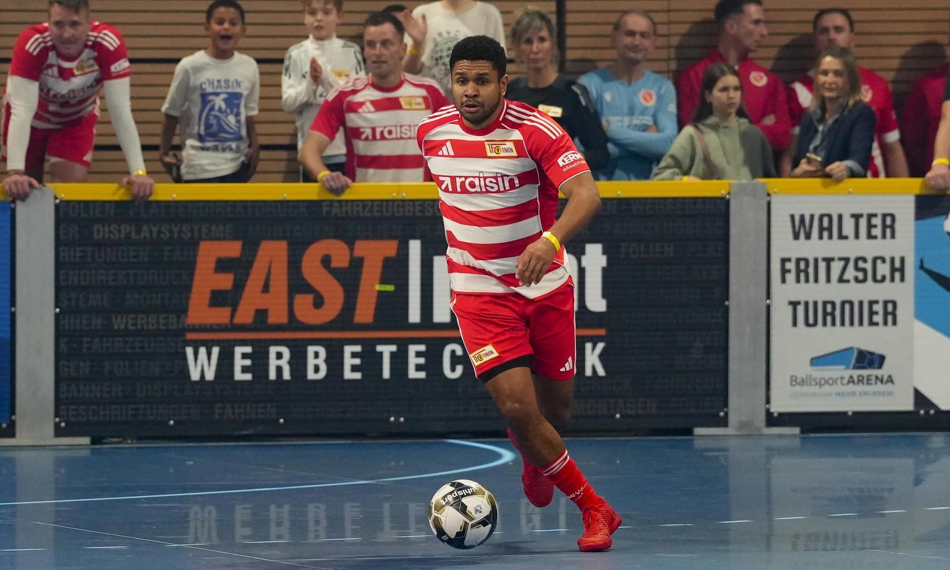 Spieler in roter und weißer Vereinsausrüstung dribbelt den Ball während eines Hallenfußballturniers. Zuschauer im Hintergrund.