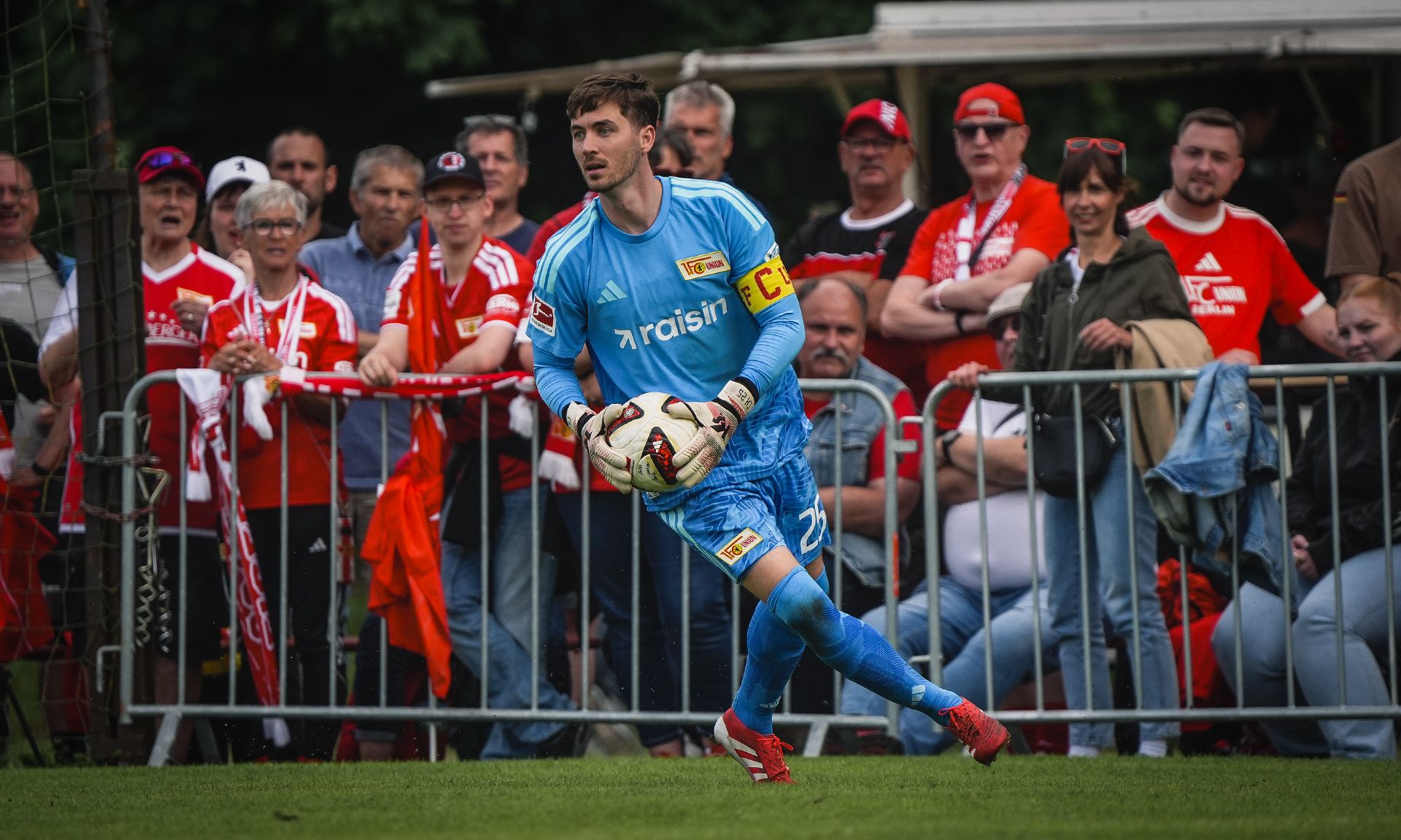 Torwart in blauer Ausrüstung hält den Ball, während eine jubelnde Menge im Hintergrund zuschaut.