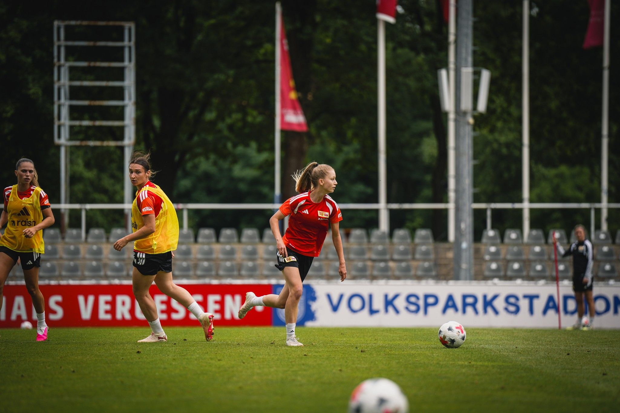 Drei Fußballspielerinnen in Trainingskleidung auf einem Spielfeld, eine Spielerin führt den Ball aus. Im Hintergrund stehen Zuschauerplätze.