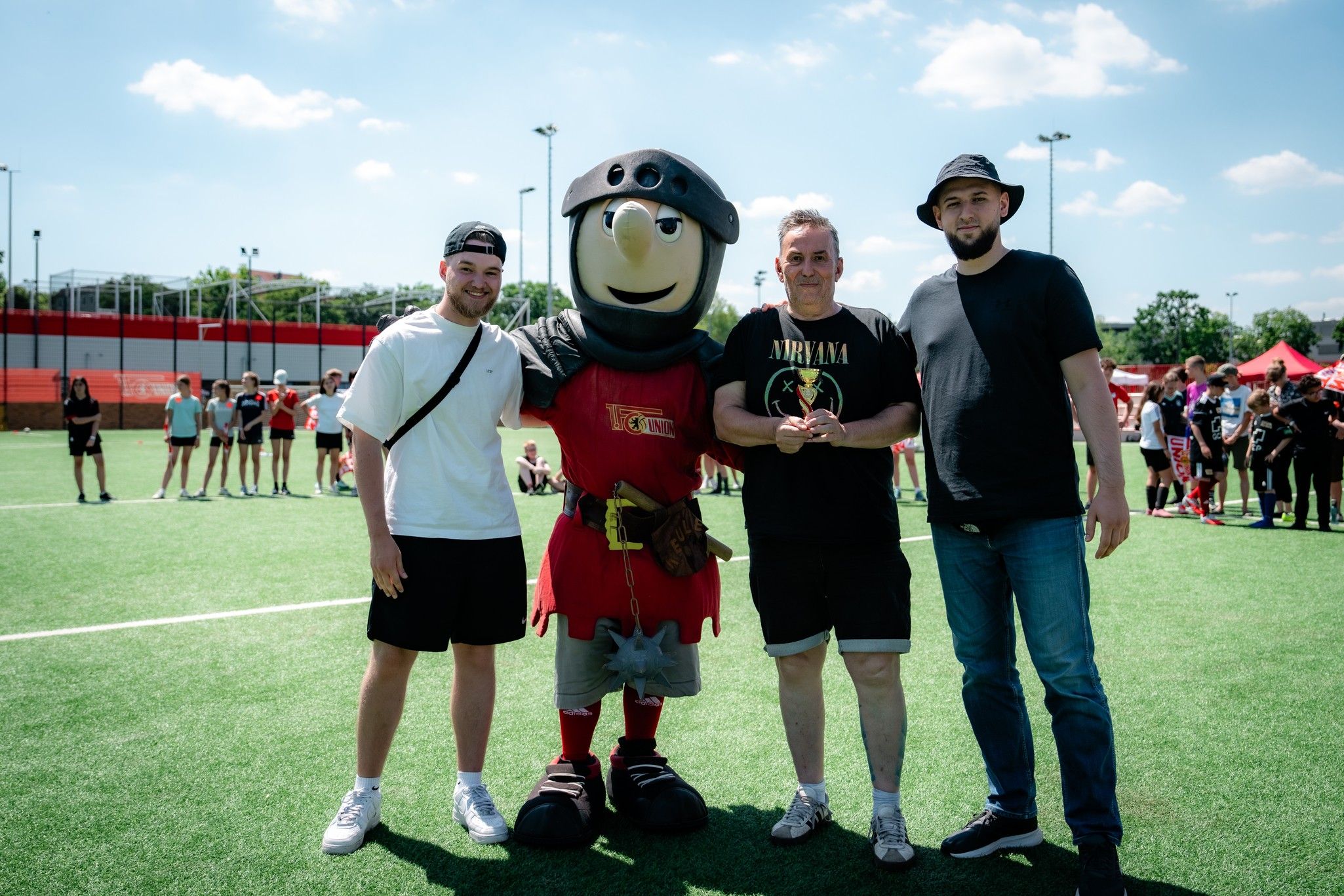 Drei Männer stehen auf einem Fußballfeld, einer trägt ein Maskottchen-Kostüm. Im Hintergrund sind weitere Personen und Stände zu sehen.