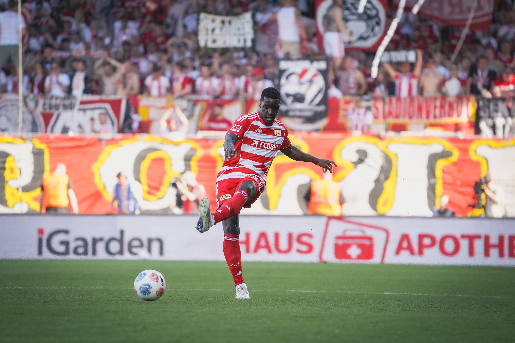 Spieler in rot-weißer Fußballausrüstung tritt den Ball vor einer jubelnden Menge im Stadion.