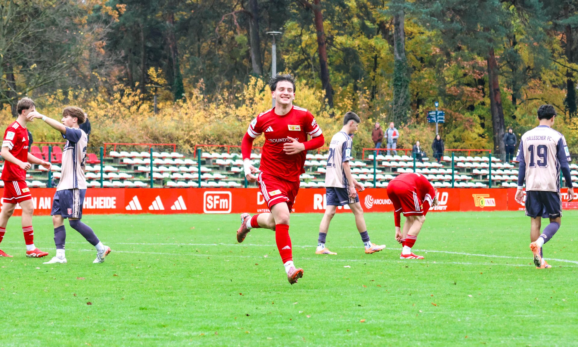 Ein junger Fußballspieler in rotem Trikot lächelt und läuft auf einem Sportplatz, während andere Spieler im Hintergrund aktiv sind.