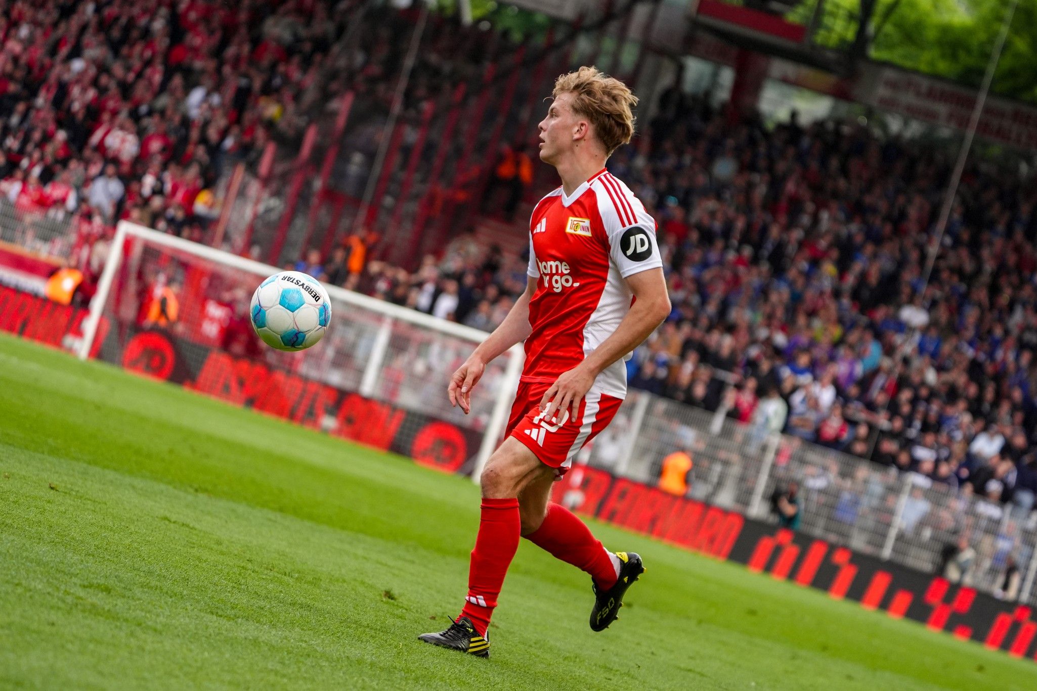 Ein Fußballspieler in rotem Trikot mit dem Ball am Fuß, vor jubelnden Zuschauern im Stadion.