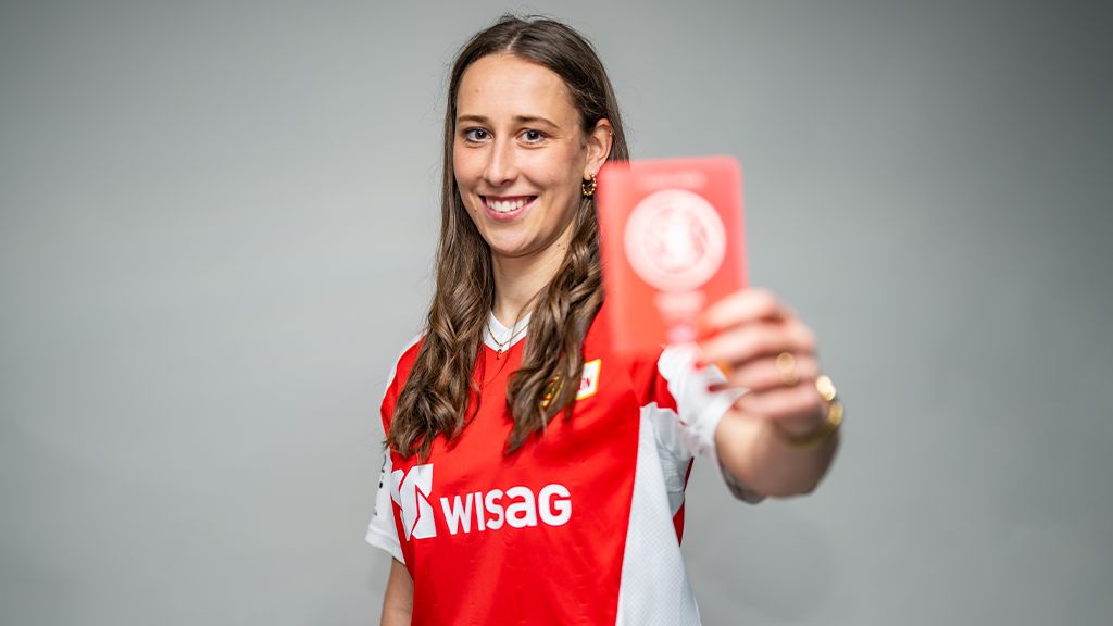 Eine Person in einem roten Trikot hält eine rote Karte in die Kamera und lächelt. Hintergrund ist grau.