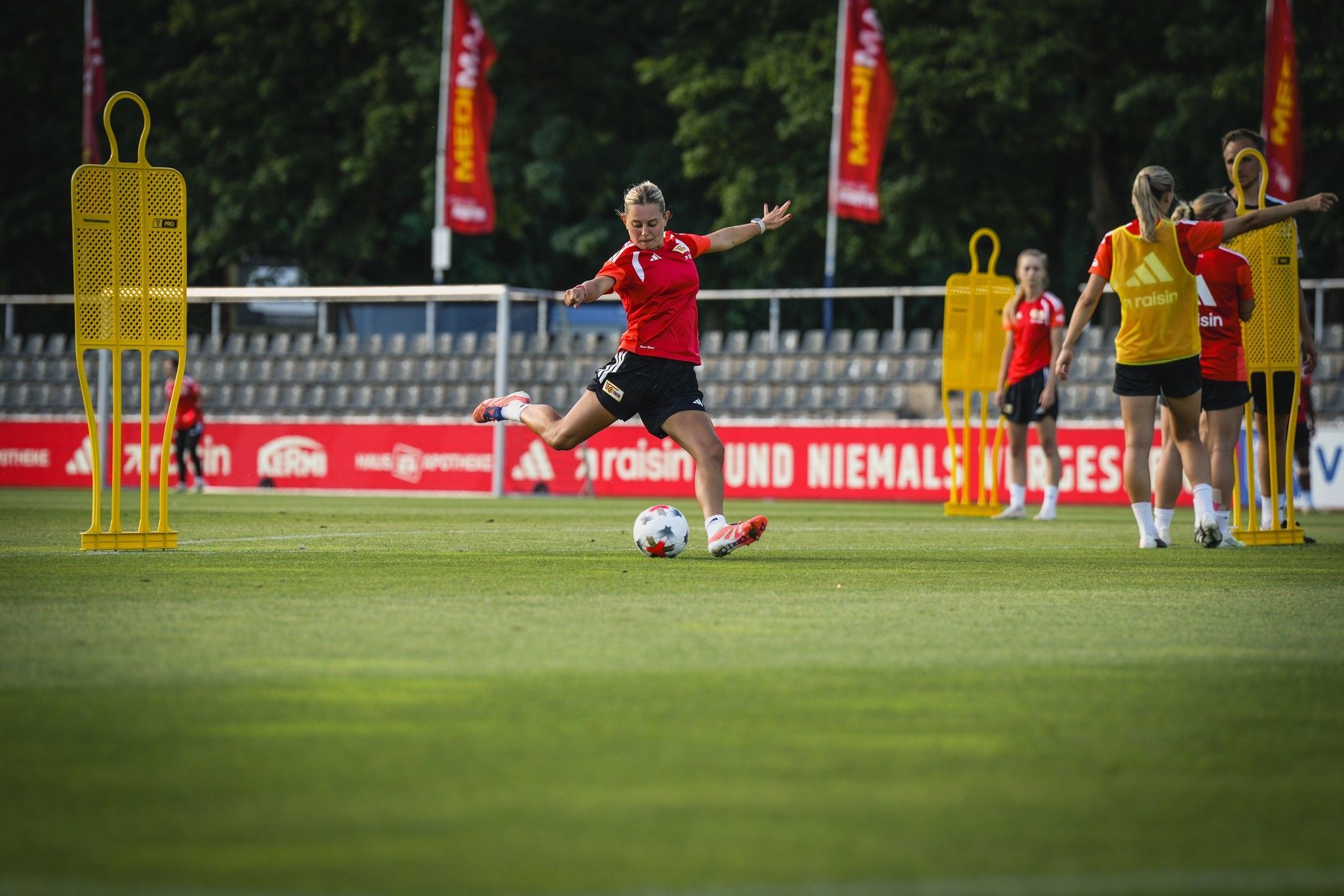 Eine Fußballspielerin trainiert und schießt den Ball auf einem Grünfeld, umgeben von Trainingshilfen und anderen Spielern.