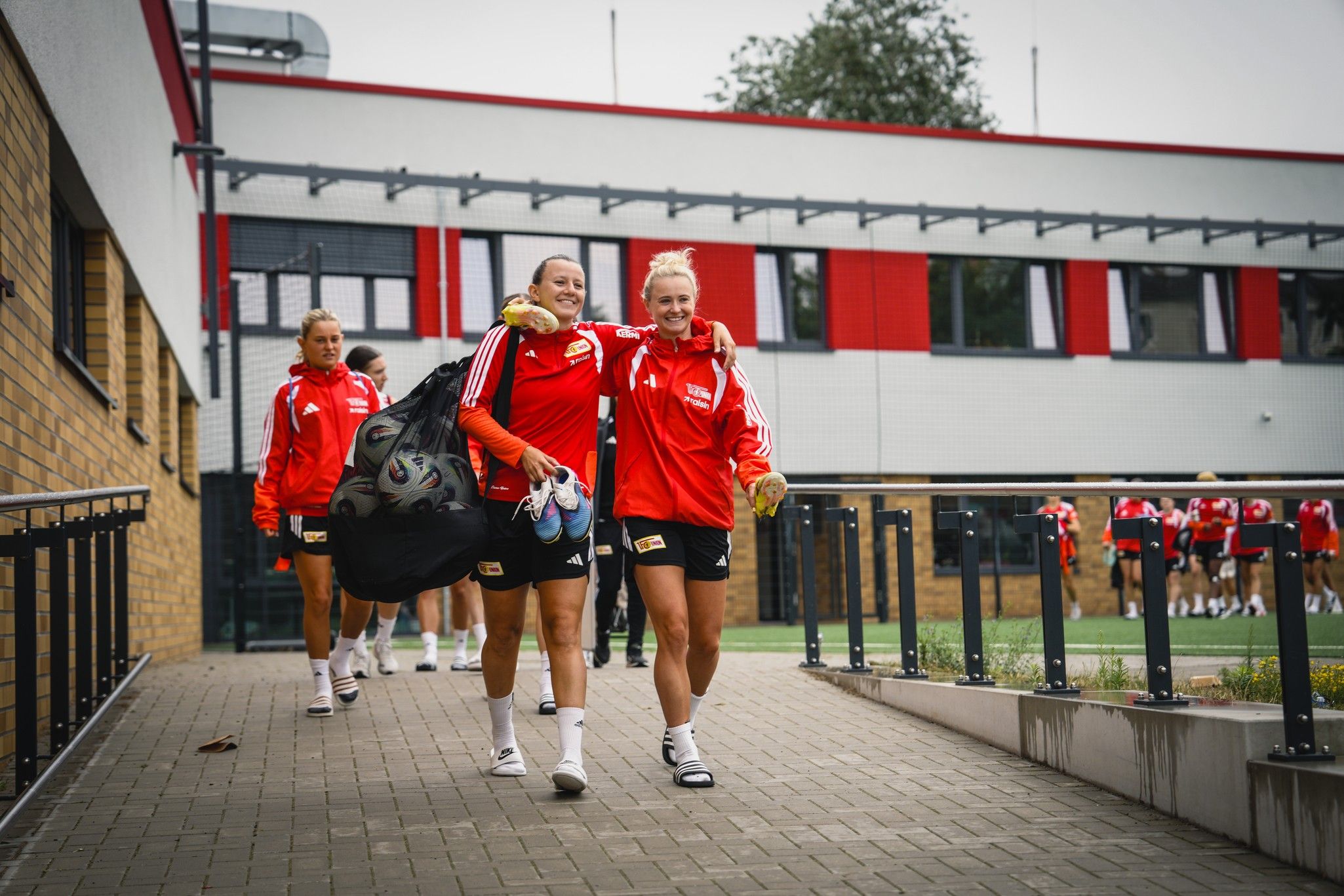 Zwei Fußballspielerinnen in roten Trainingsanzügen gehen fröhlich auf einem Weg, während weitere Spielerinnen im Hintergrund folgen.