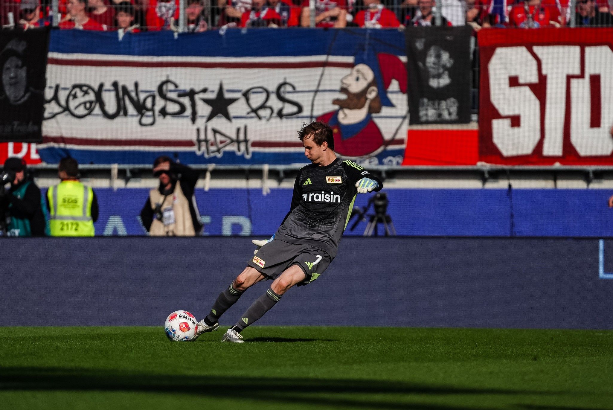 Torwart in grauer Uniform führt einen Abschlag aus, im Hintergrund sind Fans und Banner zu sehen.