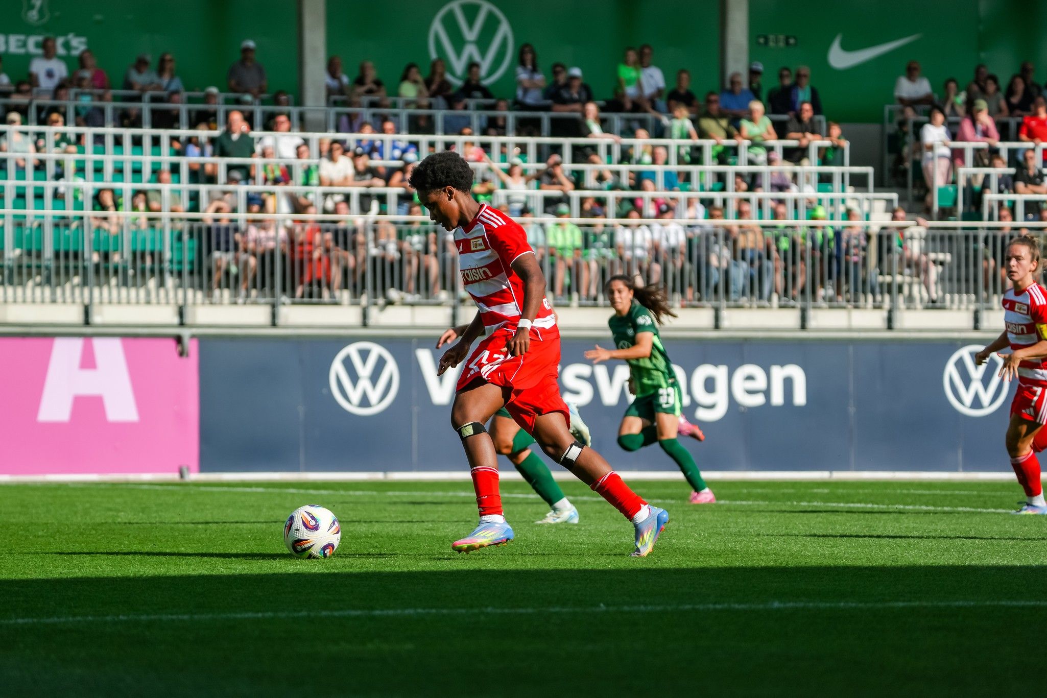 Ein Fußballspieler in rot-weißer Ausrüstung dribbelt den Ball auf einem grünen Spielfeld, während Zuschauer im Hintergrund zuschauen.