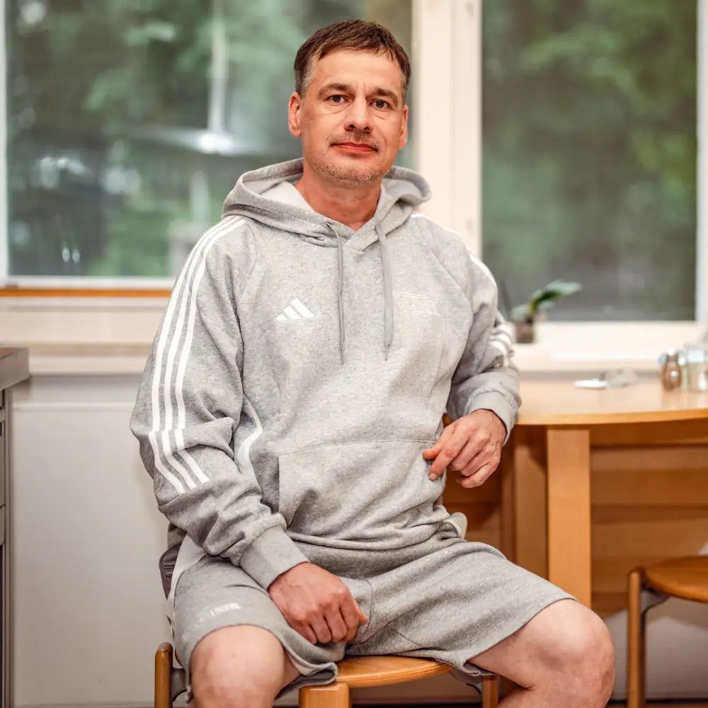 Ein Mann trägt ein graues Sportoutfit von Adidas. Er sitzt auf einem Stuhl. Der Raum ist hell. Es gibt Fenster und Holzdekoration.