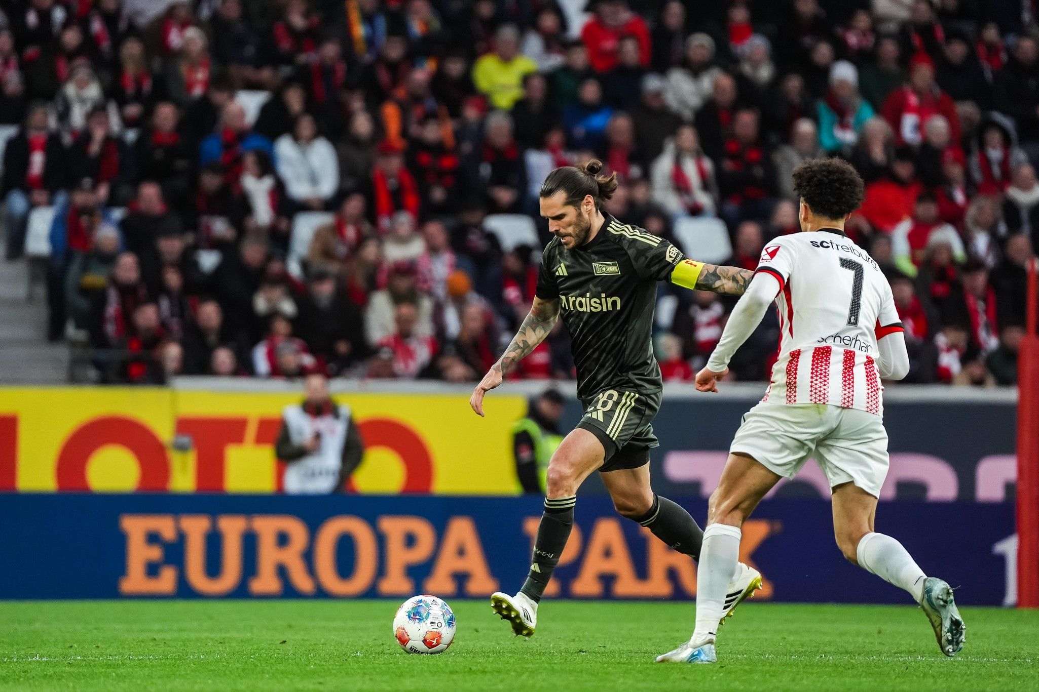Ein Fußballspieler dribbelt den Ball, während ein Gegner ihm nachläuft. Im Hintergrund ist ein Publikum im Stadion zu sehen.