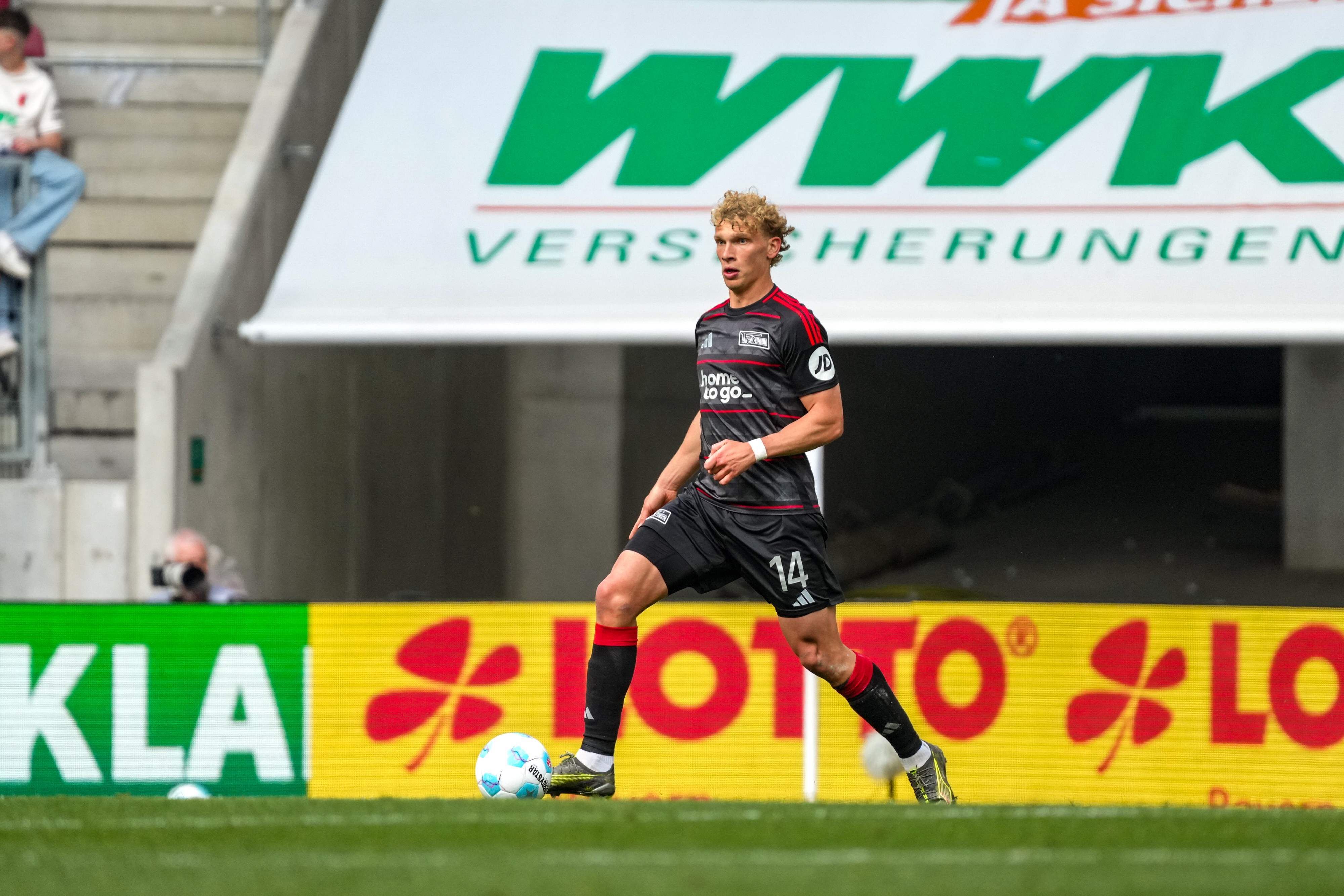 Ein Fußballspieler mit der Nummer 14 dribbelt den Ball auf dem Feld, umgeben von Werbebannern.