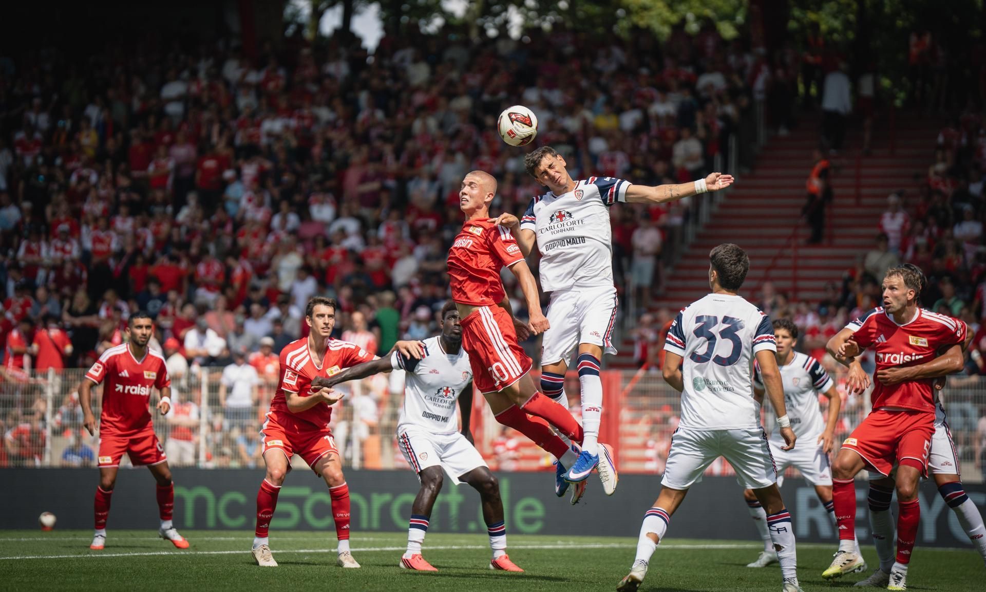 Zwei Fußballspieler springen im Luftduell, während die Zuschauer im Hintergrund ein spannendes Spiel verfolgen.