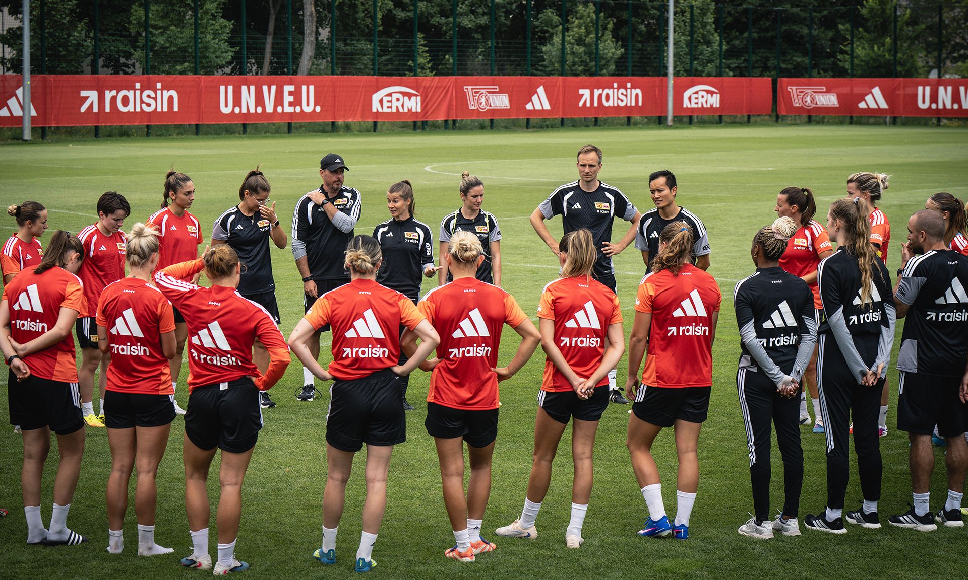 Frauenfußballmannschaft versammelt sich auf einem Trainingsplatz, während Trainer Anweisungen gibt. Spielerinnen in Trainingskleidung.