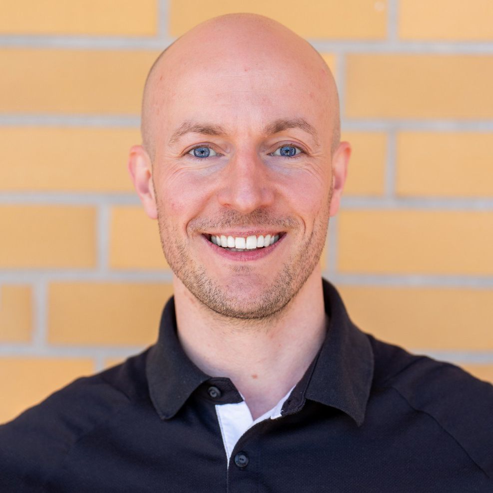 Lächelnder Mann mit kahlem Kopf und blauen Augen, trägt ein schwarzes Polohemd, vor einer hellen Ziegelwand.