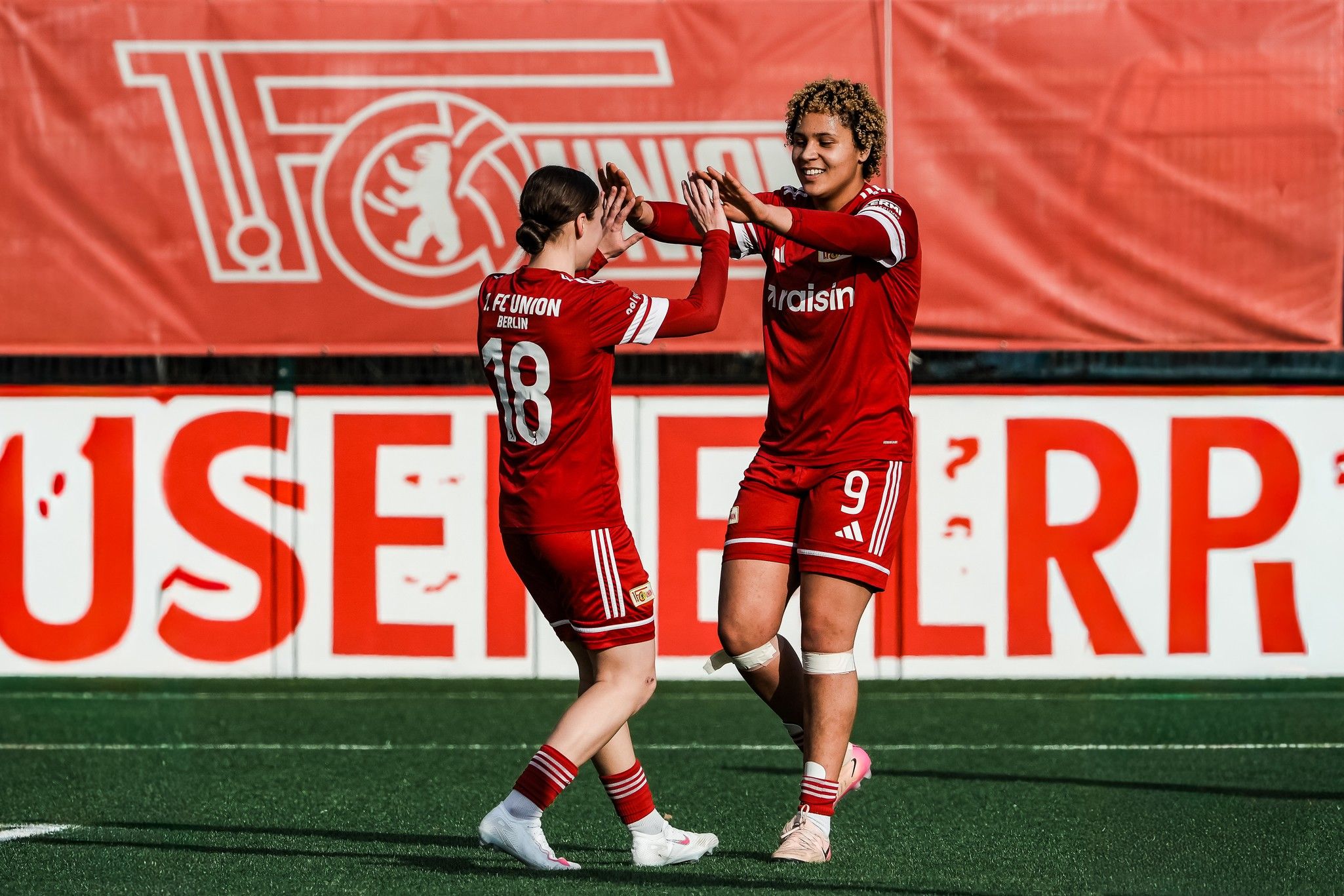 Zwei Fußballspielerinnen in roten Trikots feiern einen gelungenen Spielzug auf dem Feld. Im Hintergrund ein Banner.