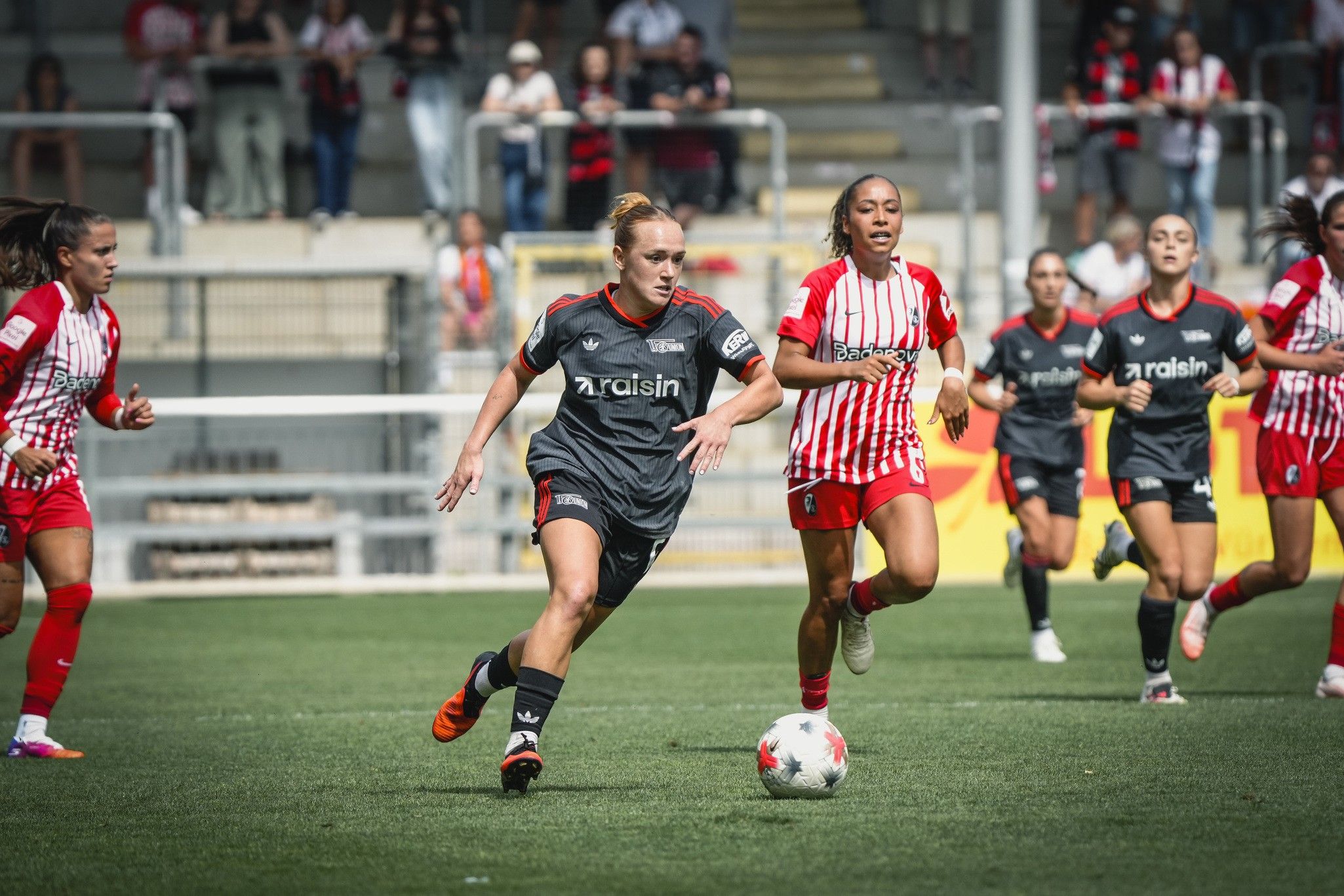 Zwei Fußballspielerinnen in actionreichen roten und schwarzen Trikots auf dem Spielfeld, im Hintergrund Zuschauer auf der Tribüne.