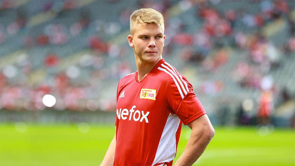 Ein junger Fußballspieler in rotem Trikot mit dem Schriftzug "Union" steht auf dem Spielfeld. Im Hintergrund sind Zuschauertribünen sichtbar.