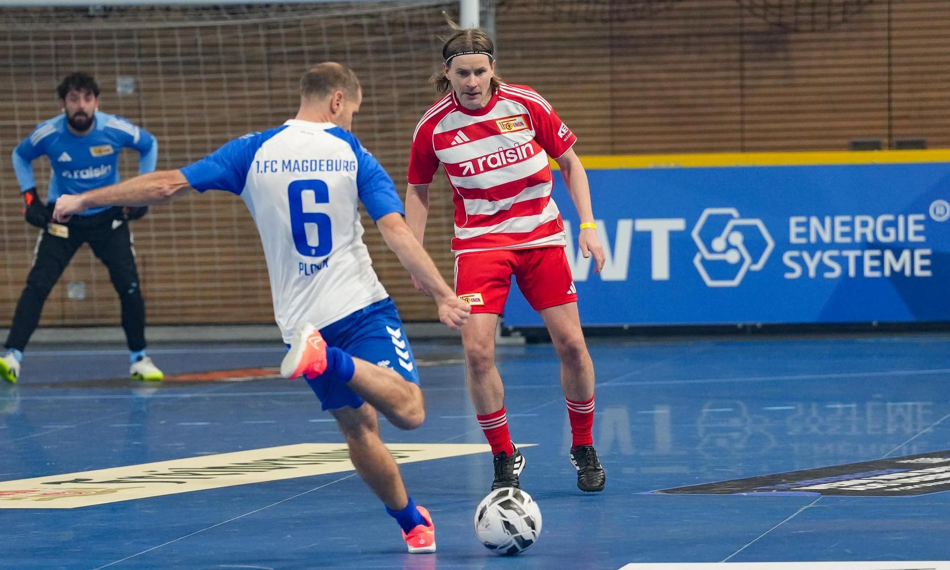 Ein Fussballspiel im Hallenformat, ein Spieler mit blauer Trikotnummer 6 schießt auf den Ball, während ein Spieler in Rot darauf reagiert.