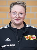 Eine lächelnde Frau mit Brille trägt eine schwarze Jacke mit dem Logo von Union Berlin. Der Hintergrund ist gemustert.