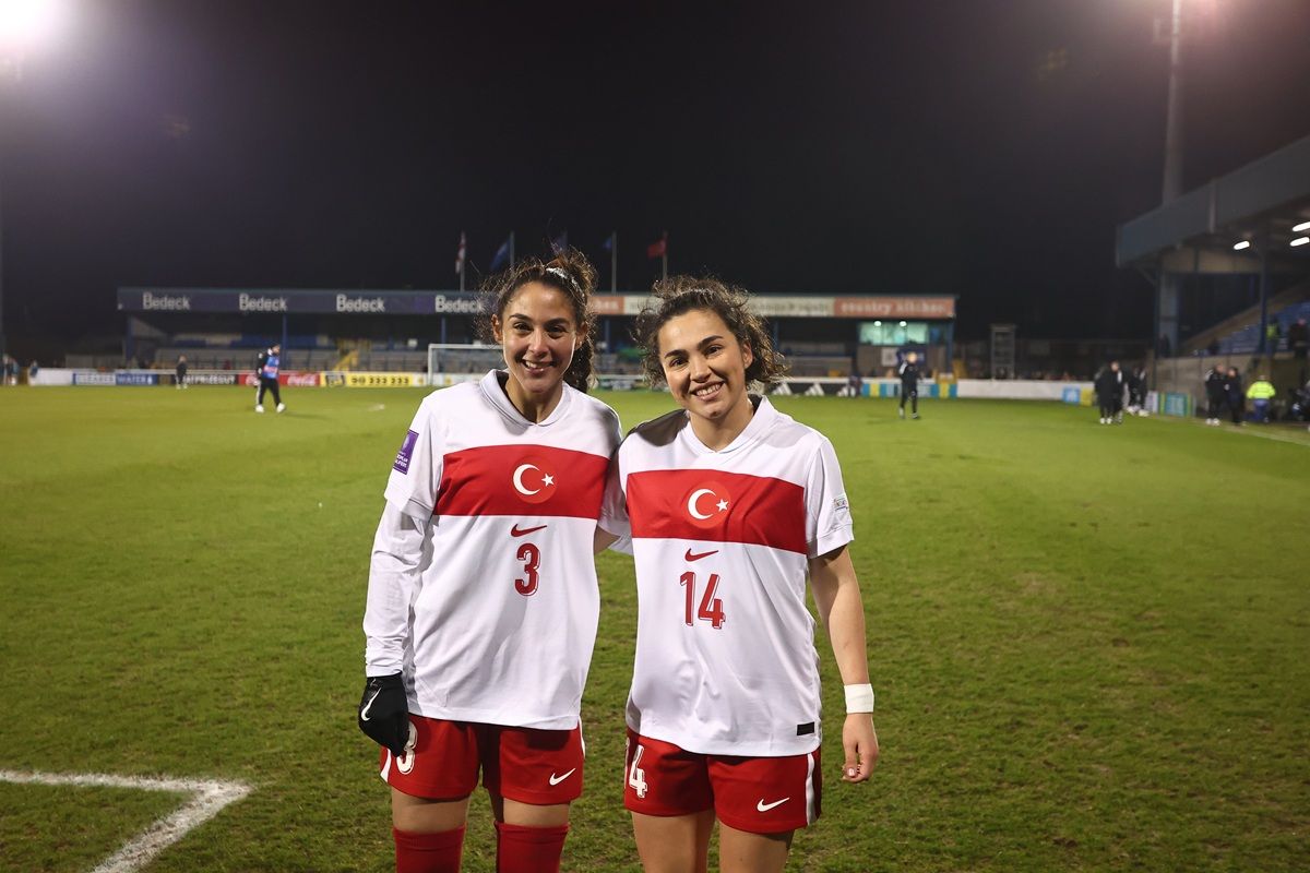 Zwei Fußballspielerinnen in weißen Trikots mit roten Akzenten und türkischen Flaggen, lächeln auf einem grünen Spielfeld.